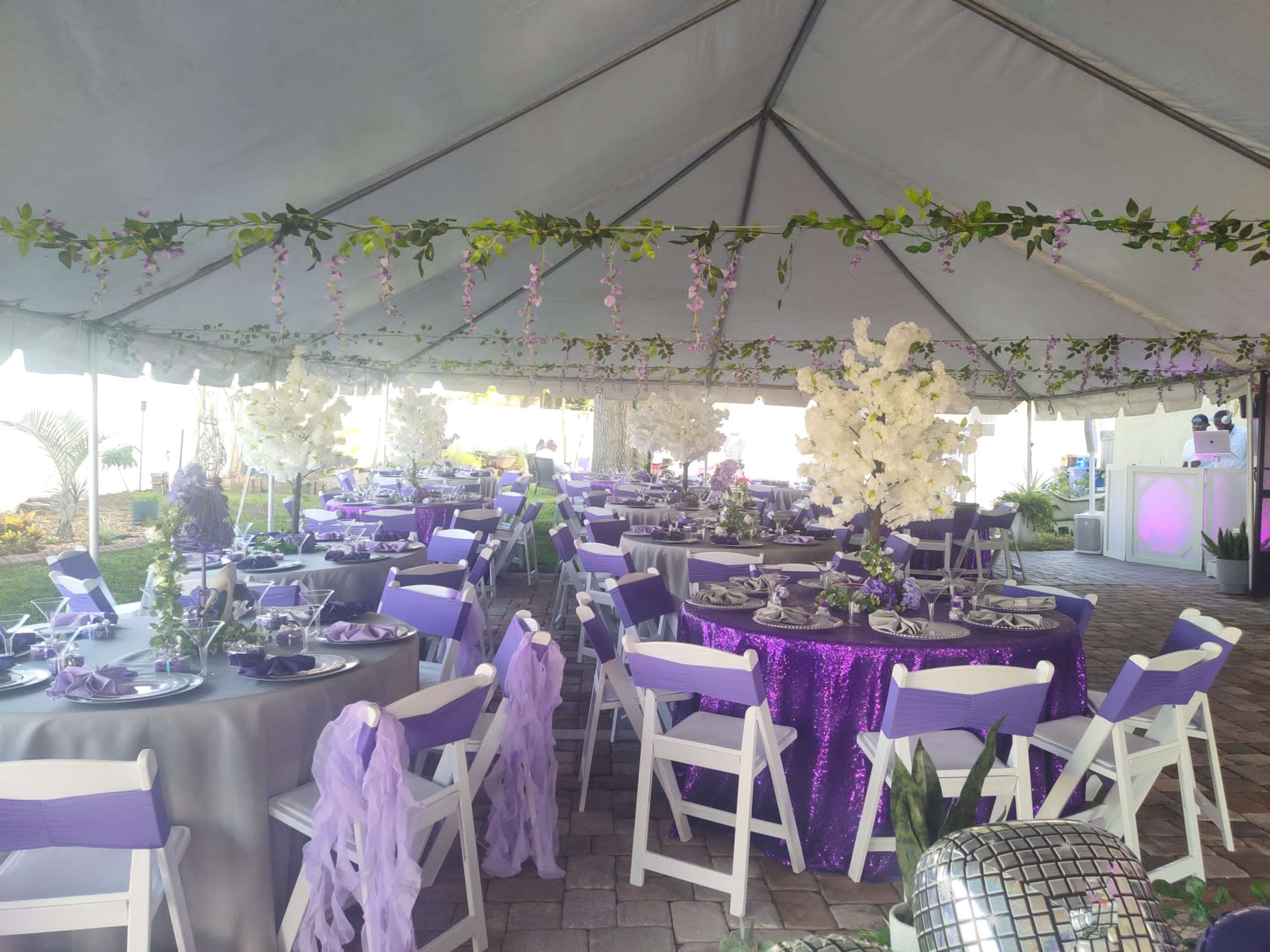 A decorated tent with round tables covered in purple and gray linens, featuring floral centerpieces and chairs adorned with fabric sashes.