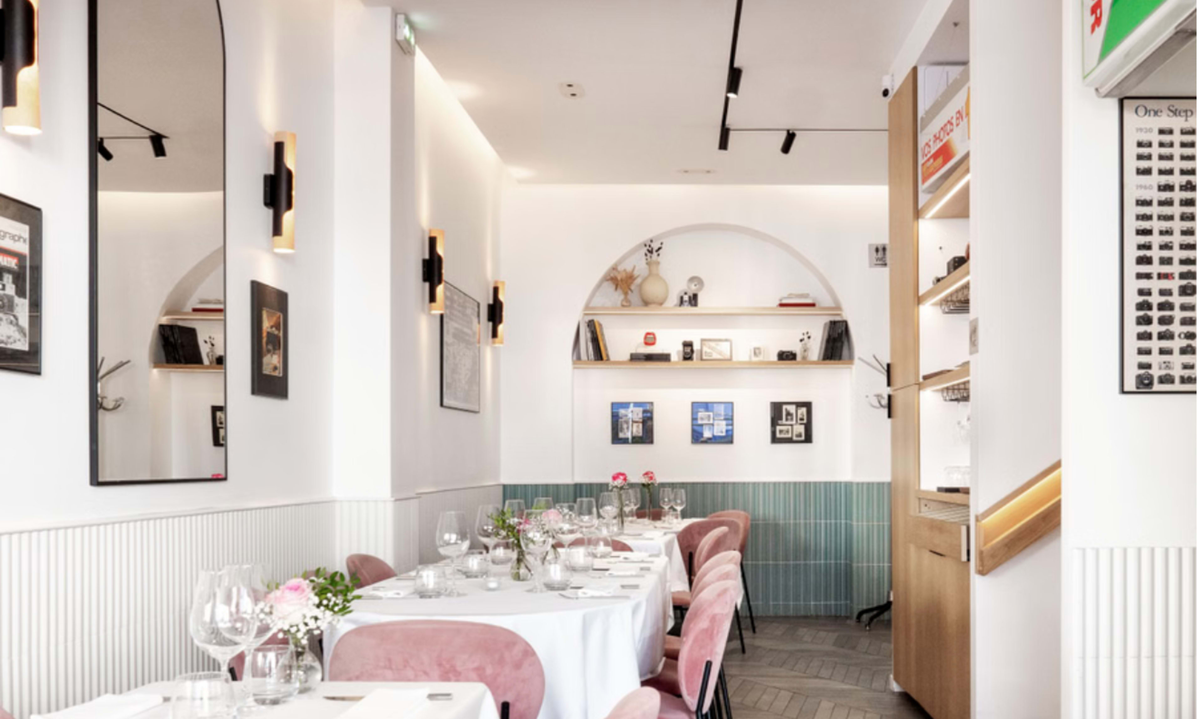 The image shows a dining area in a modern restaurant featuring white tablecloths, pink chairs, and decorative shelves along the walls.