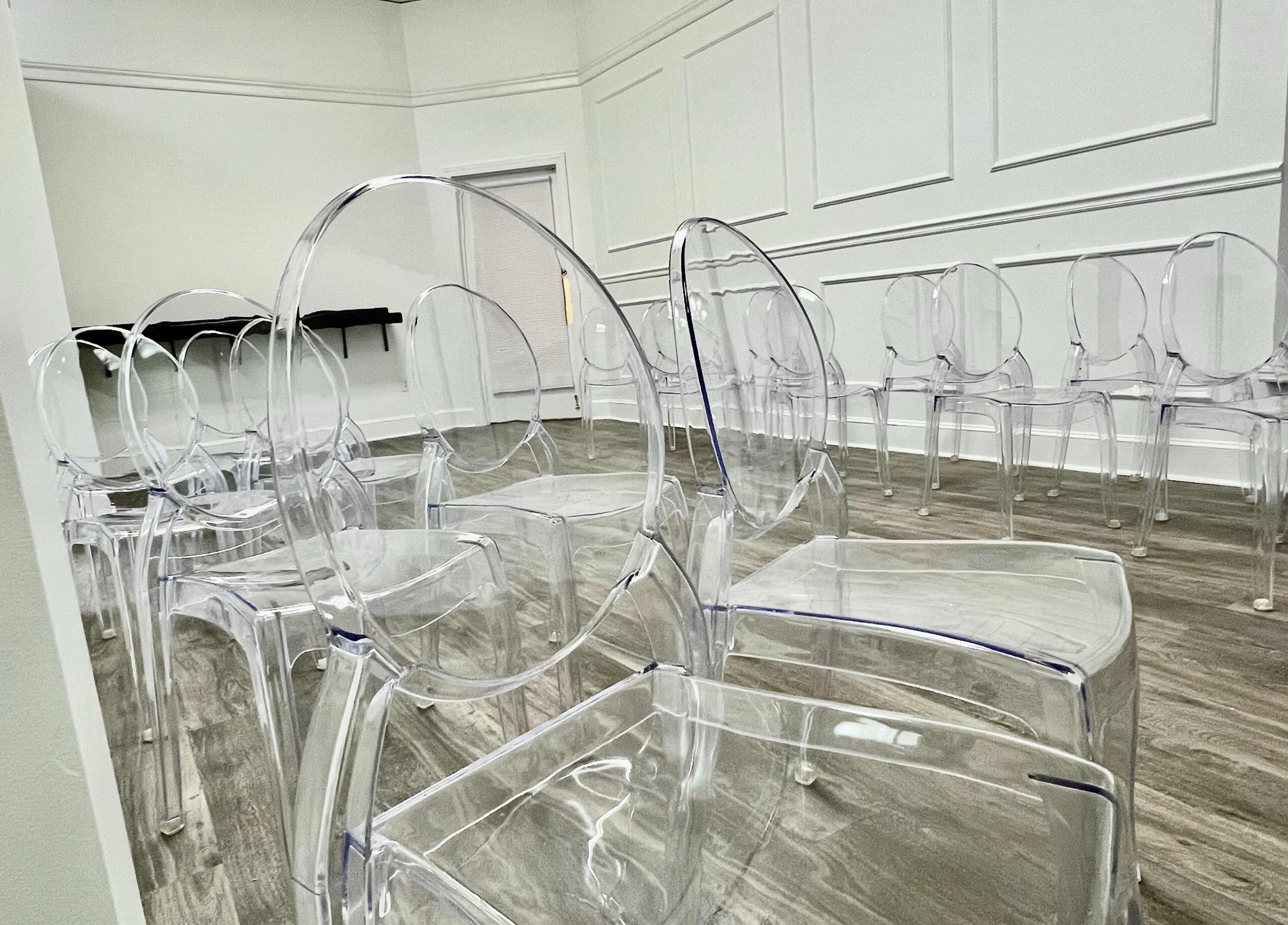 A row of transparent chairs is arranged in a minimally decorated room with white walls and a wooden floor.