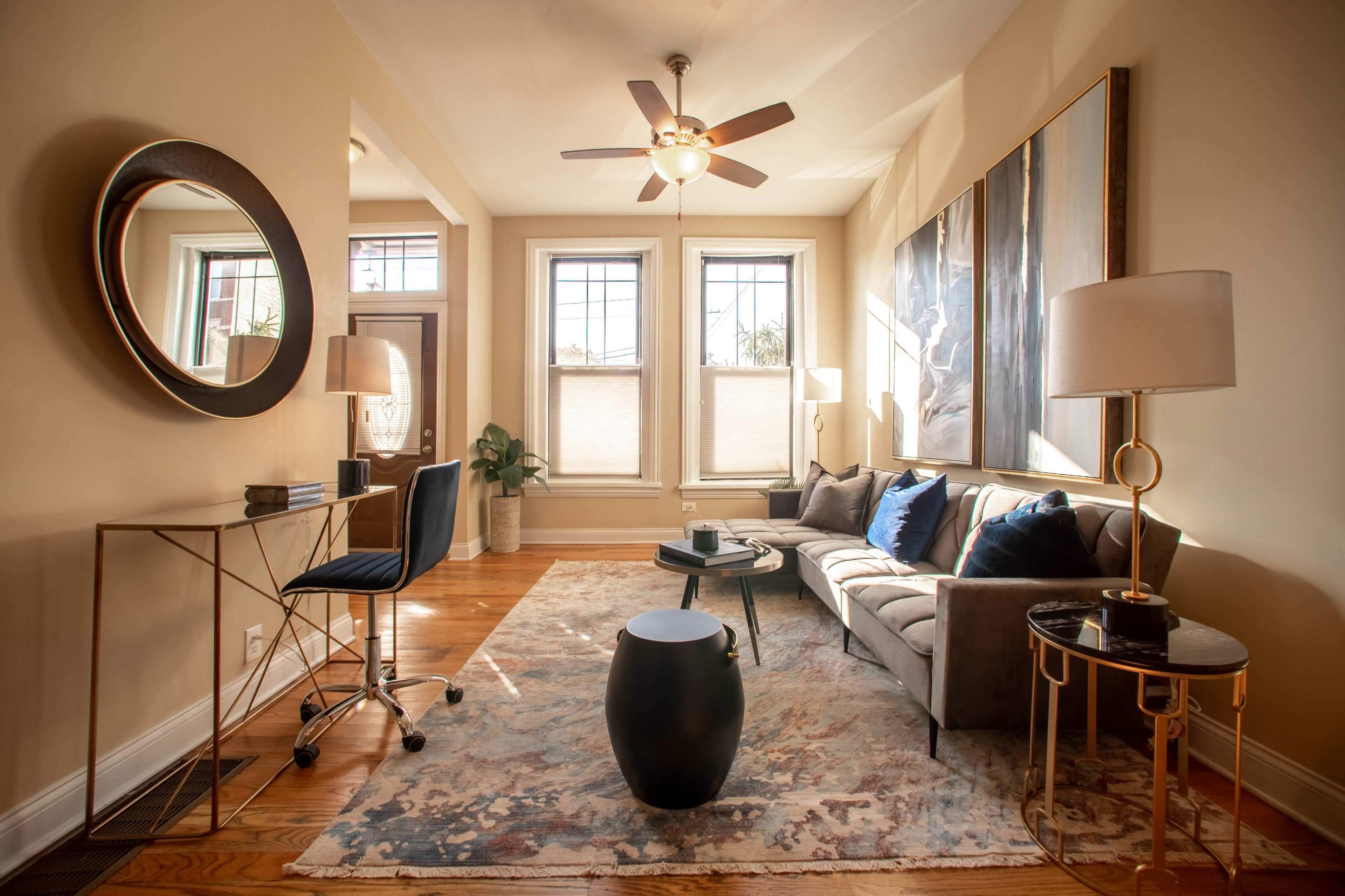 A cozy living room with a gray sofa, a round black side table, a glass-topped desk with a chair, and large windows allowing natural light to fill the space.