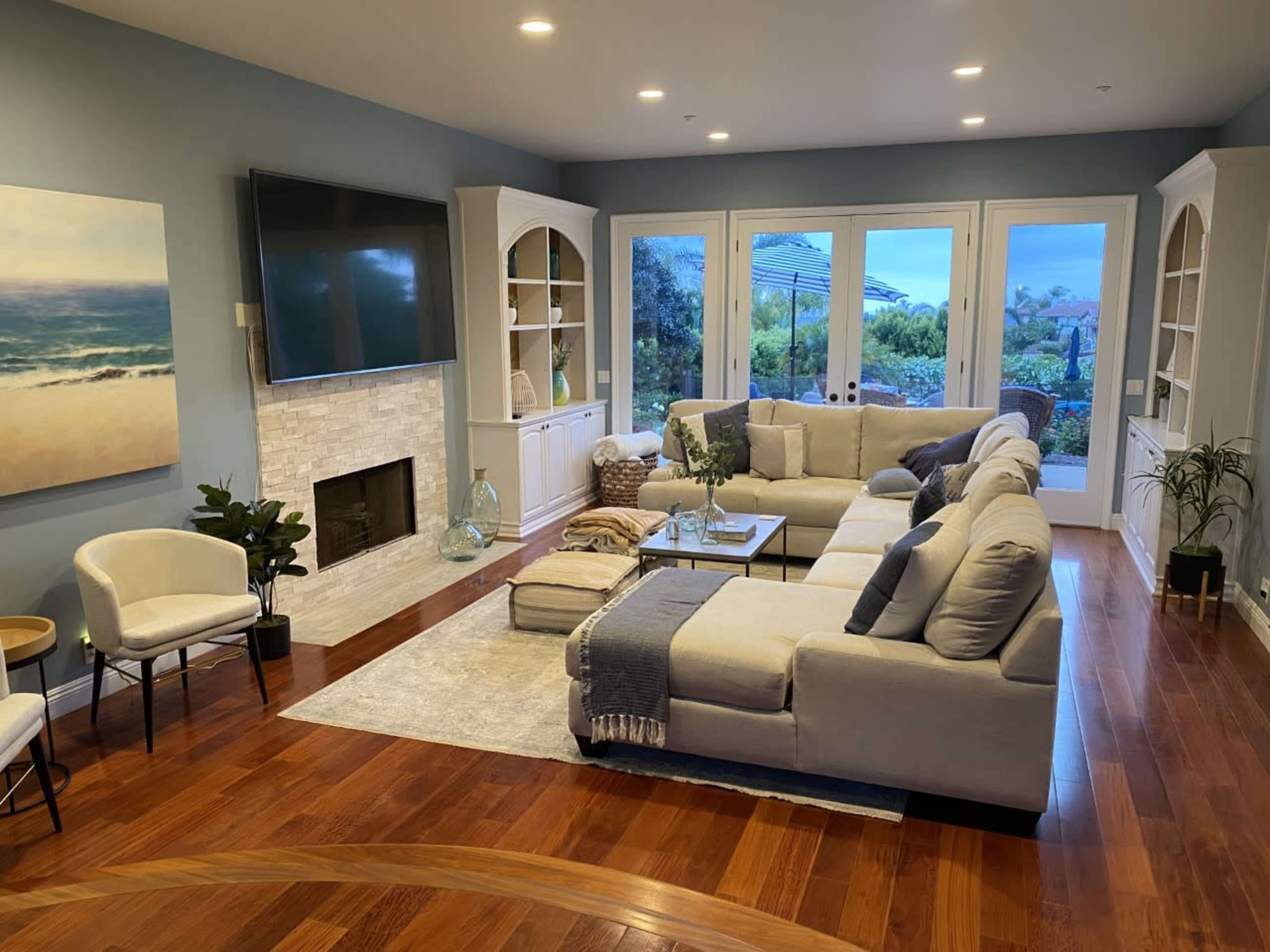 A spacious living room with a large sectional sofa, a television mounted on the wall, and glass doors leading to an outdoor view, all complemented by wooden flooring and modern decor.