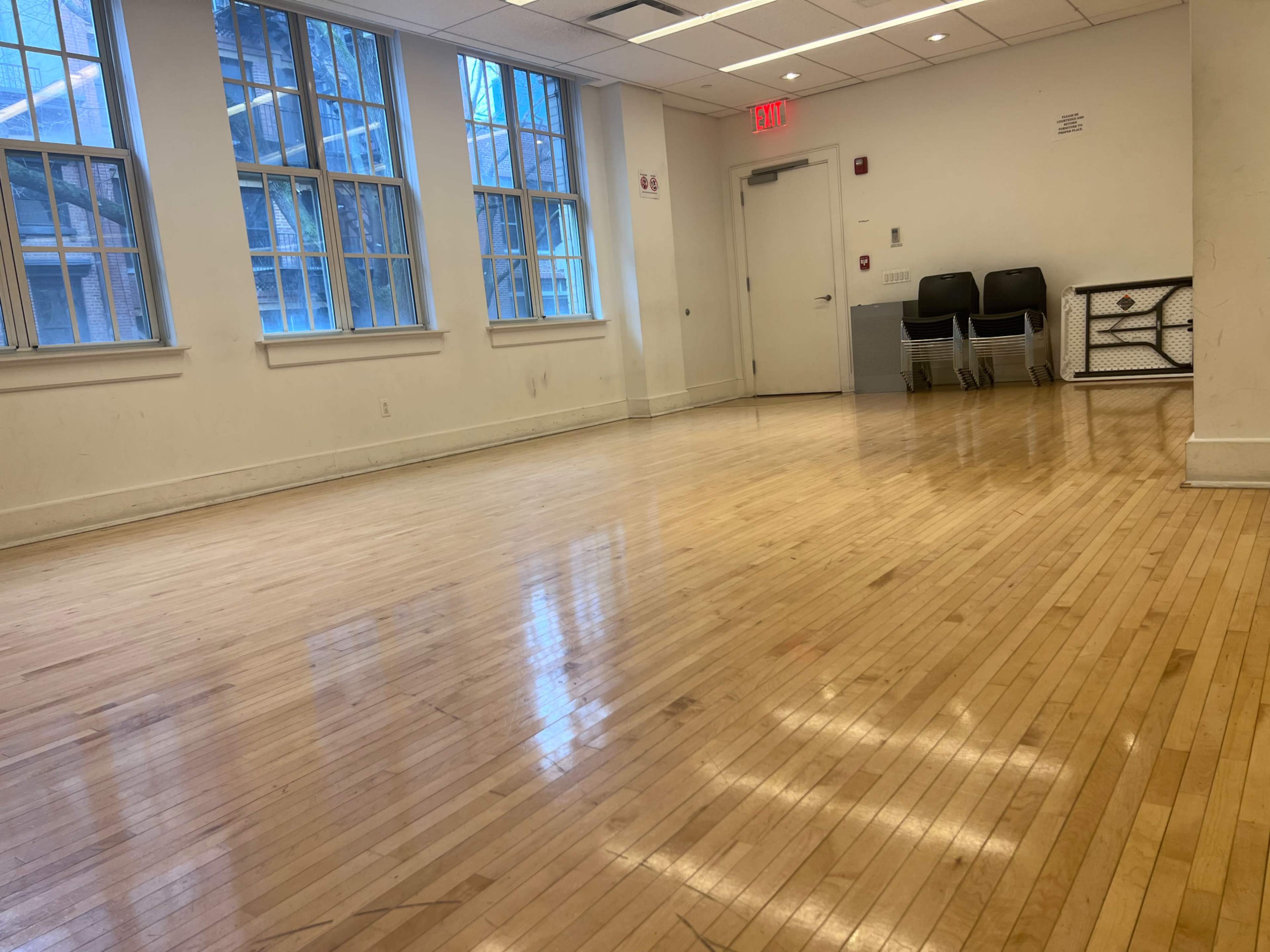 The image shows a spacious, empty room with wooden floors, large windows, and a stack of chairs against the wall.
