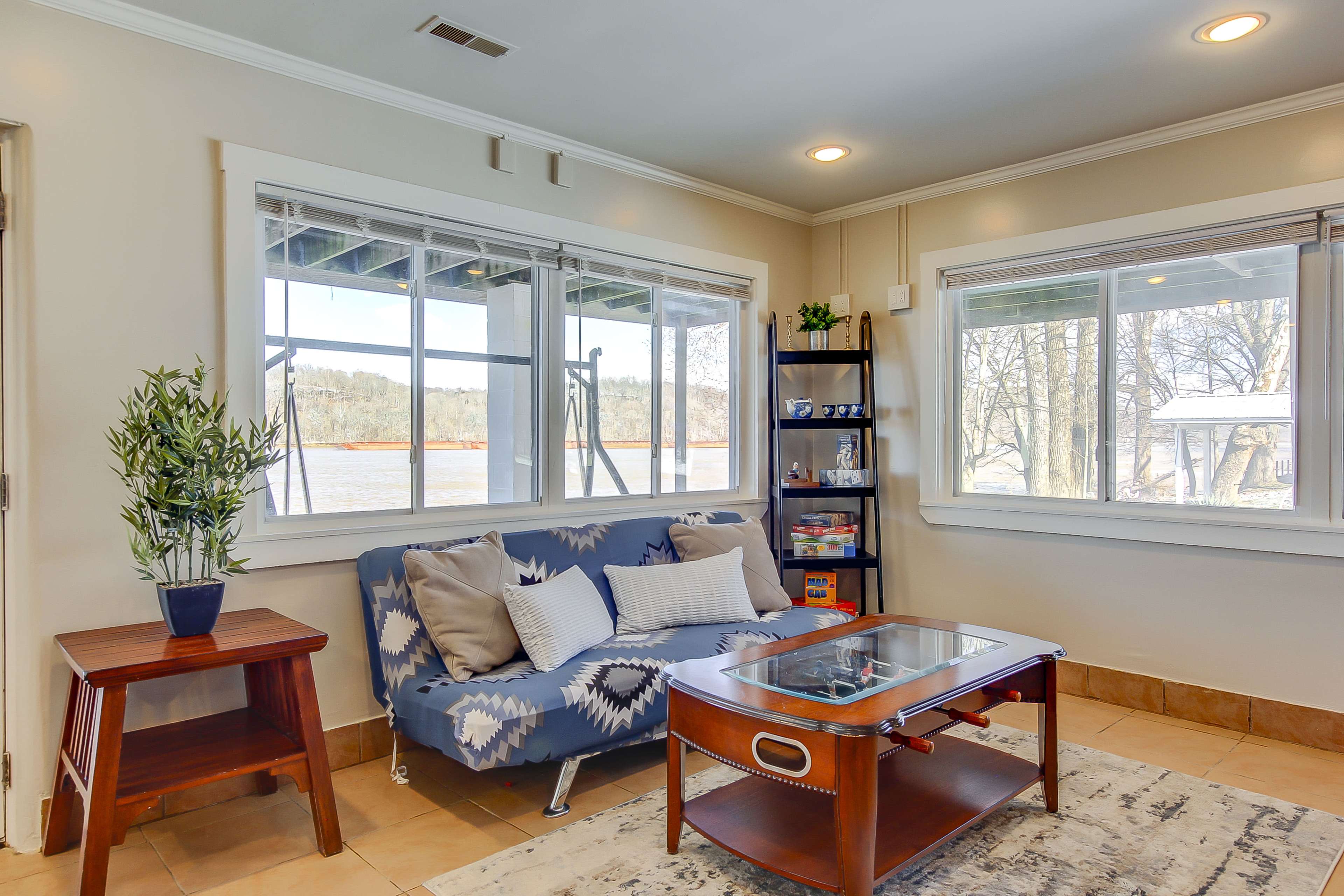 A cozy sitting area with a blue patterned sofa, a glass-top coffee table, and a bookshelf, all illuminated by natural light streaming through large windows overlooking a river.