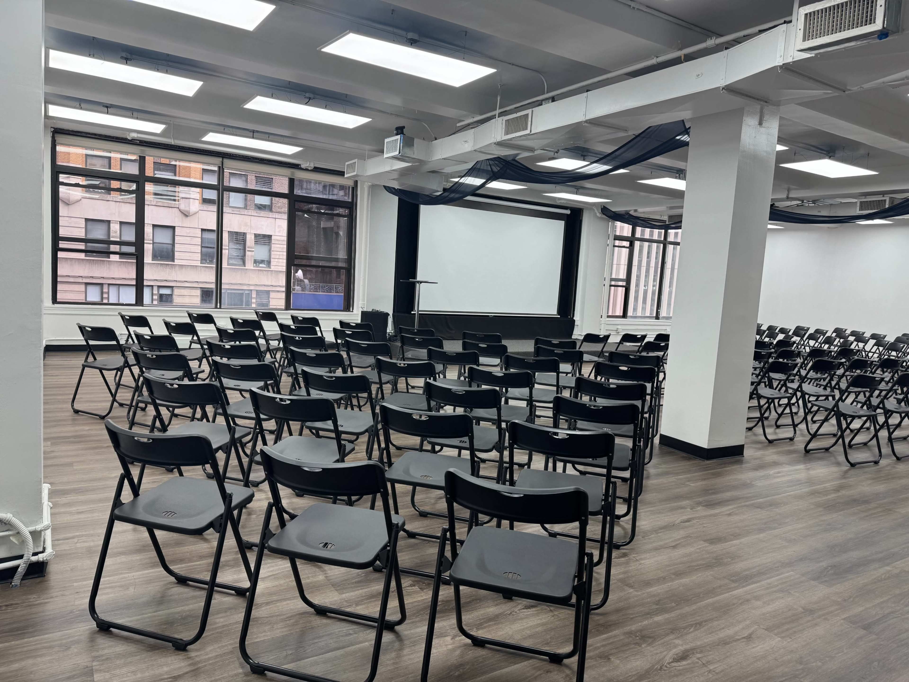 A spacious, empty room set up with rows of black folding chairs facing a large projection screen.