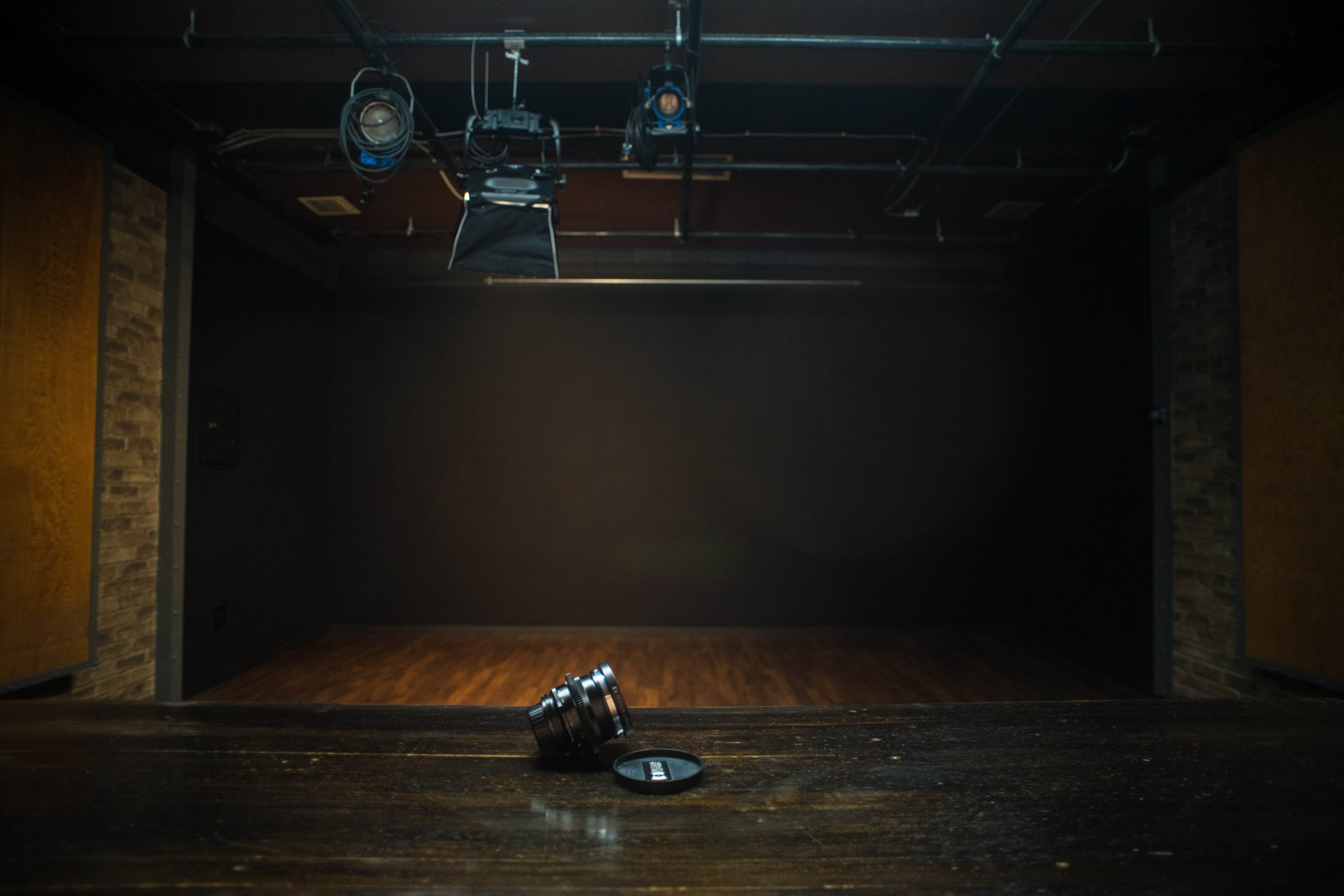 A camera lens lies on a wooden table in an empty studio space with adjustable lighting.