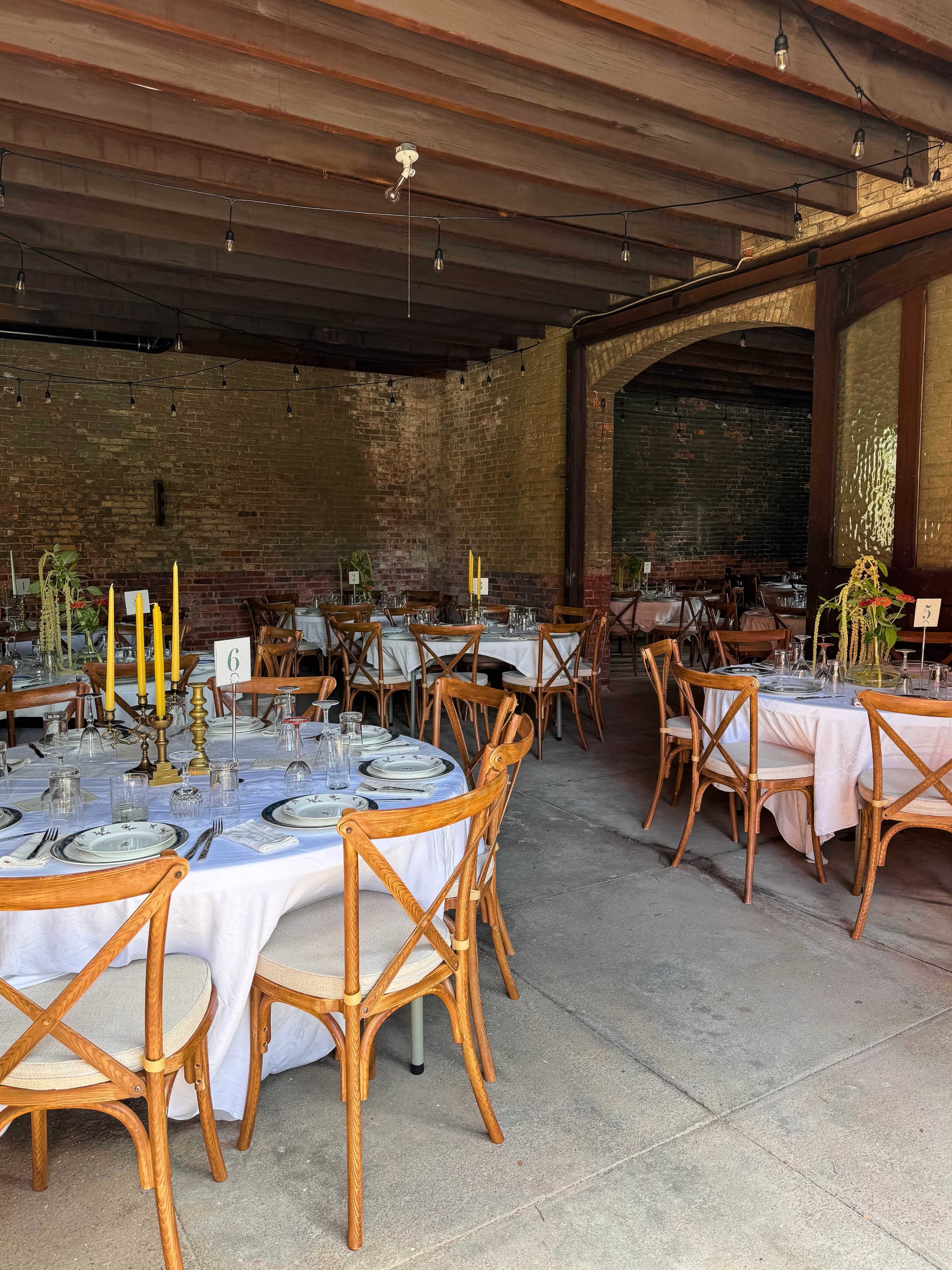 The image shows a dining area with several round tables set up with white tablecloths, wooden chairs, and decorative centerpieces, all under a wooden ceiling with exposed beams.