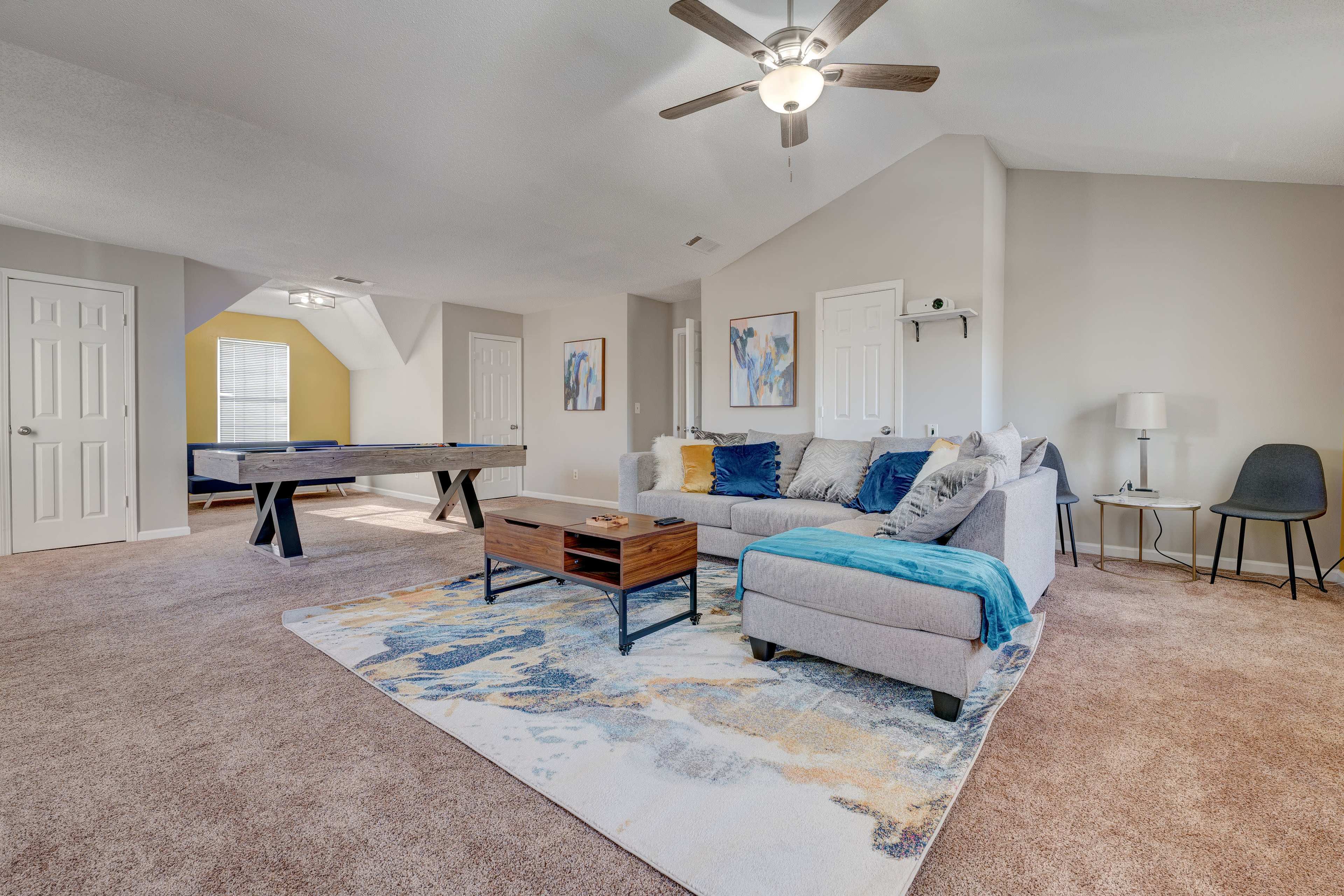A spacious living area featuring a gray sectional sofa, a coffee table, a patterned rug, and a pool table in the background.