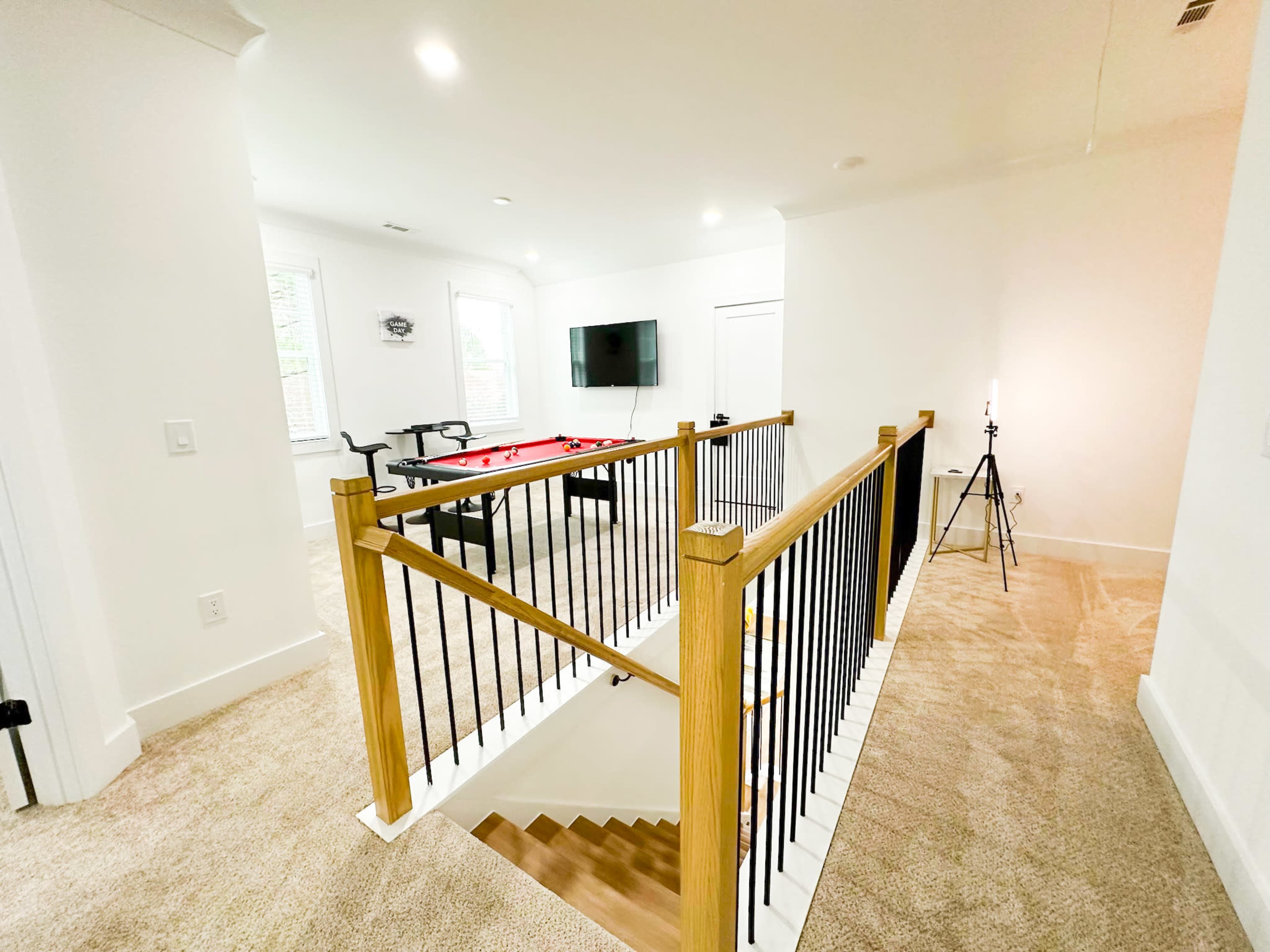 A well-lit game room features a pool table and a television, with a staircase leading down to it, surrounded by a wooden railing.