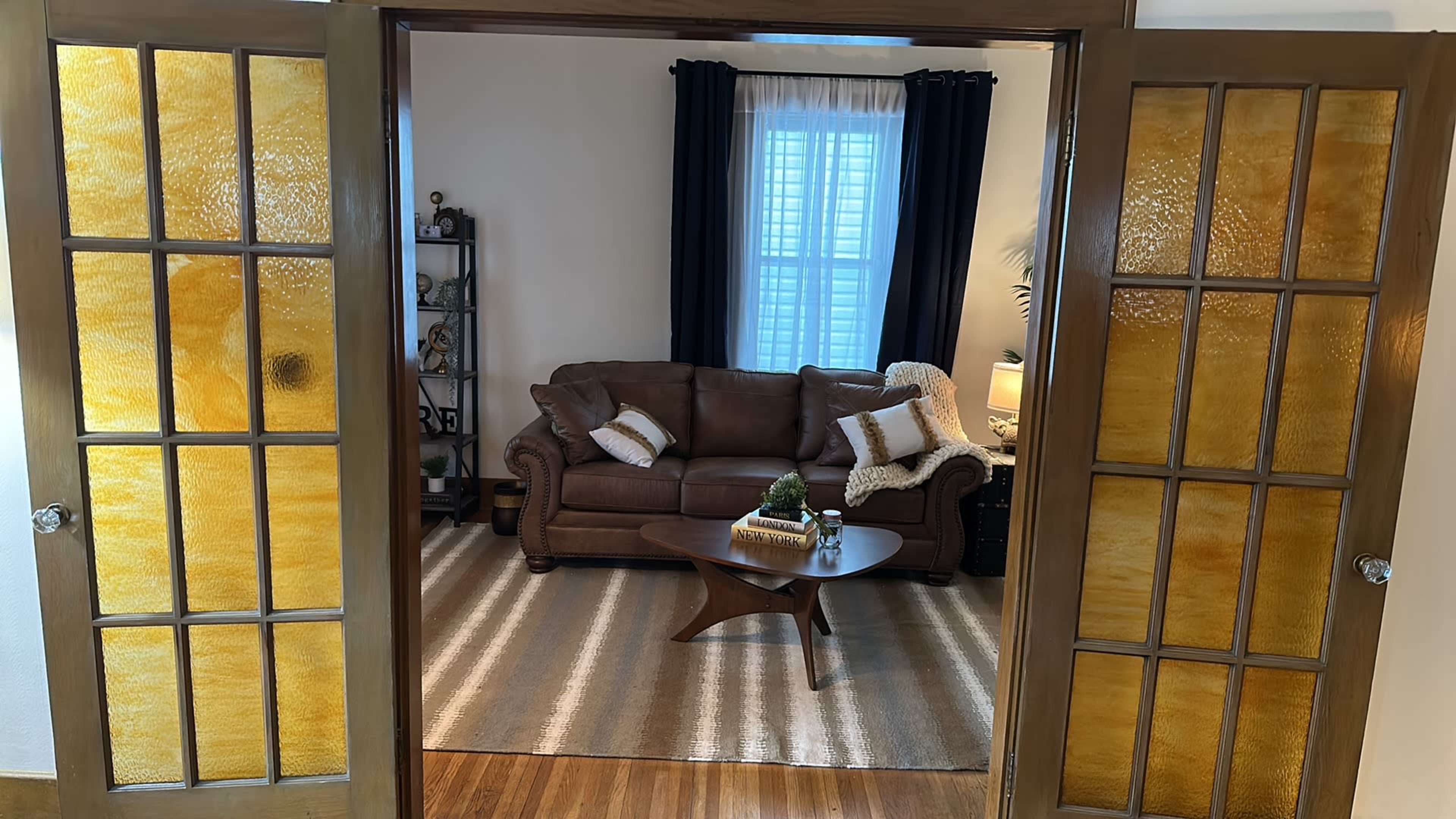 A living room with a brown leather sofa, a round coffee table, and a patterned rug, viewed through double doors with frosted glass panels.