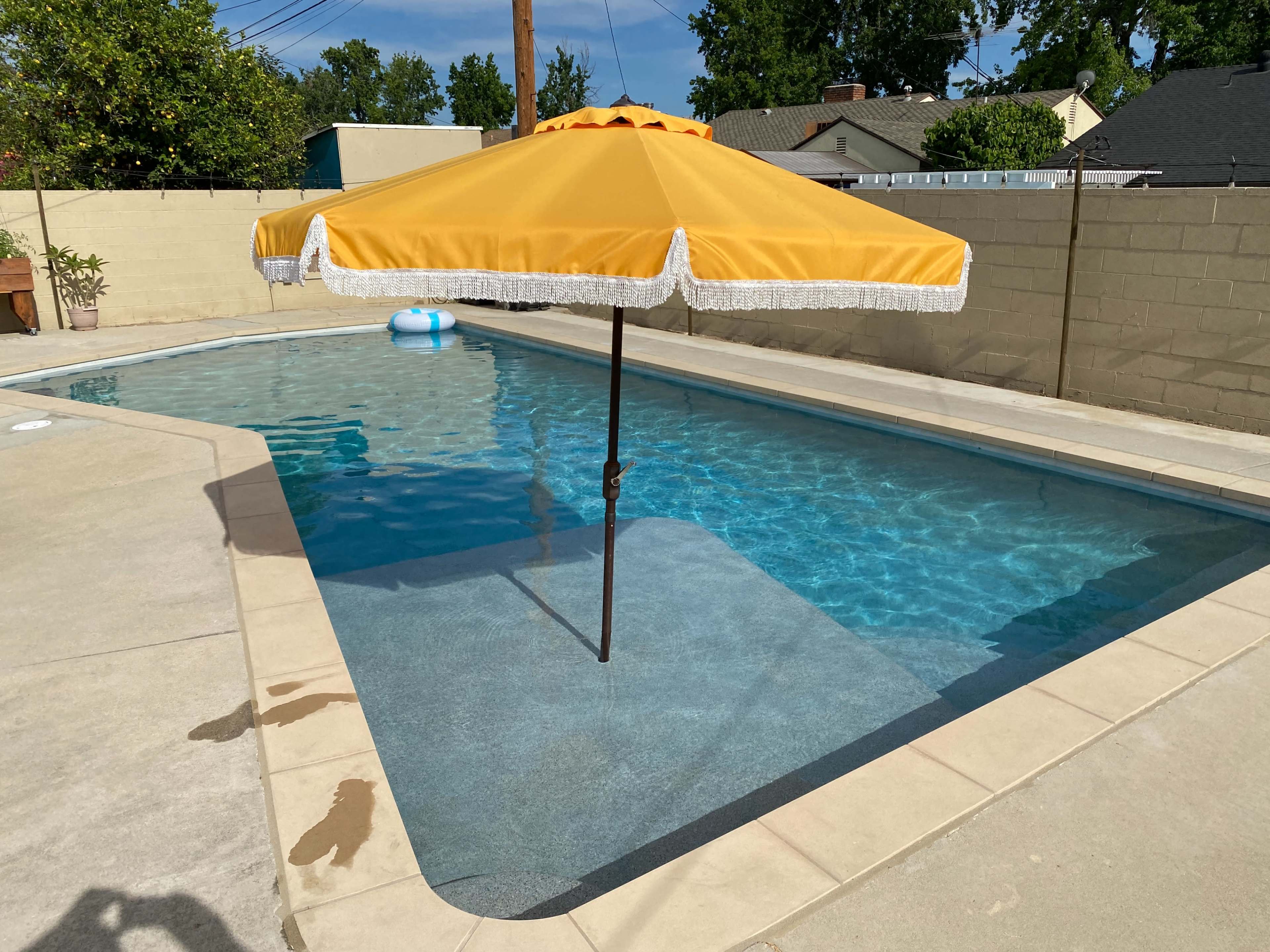 A yellow umbrella stands over a swimming pool with clear blue water, surrounded by a concrete deck and a beige wall.