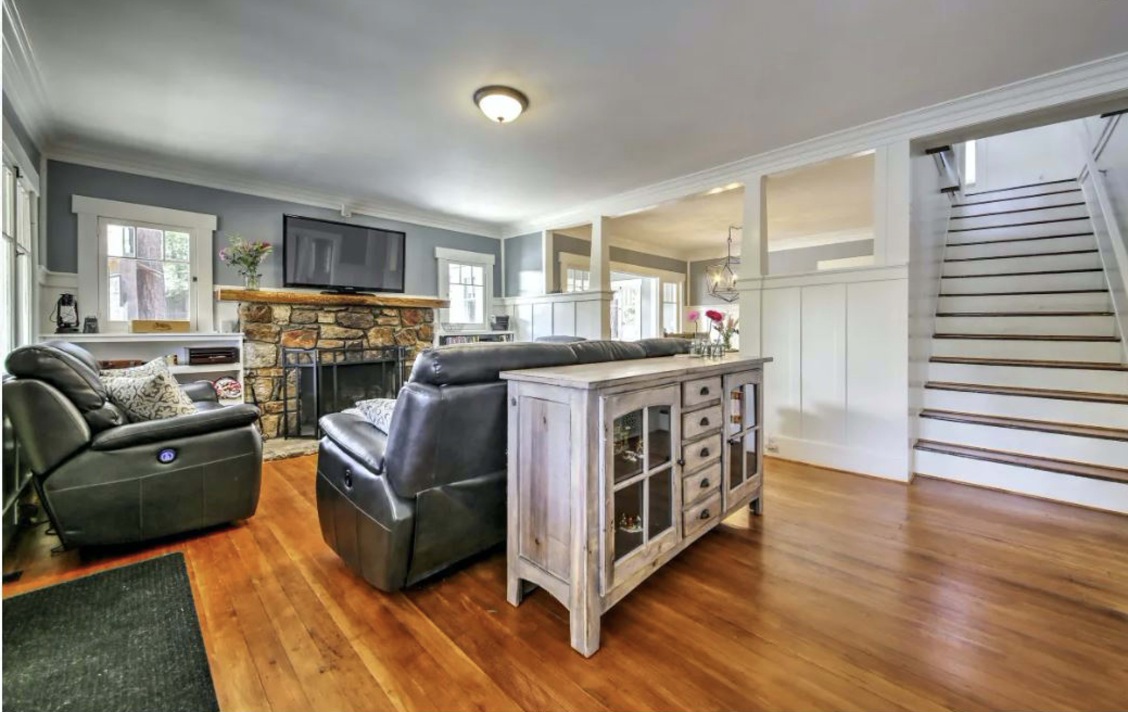 A living room with a stone fireplace, television mounted on the wall, and a wooden side table, alongside a staircase leading to an upper level.