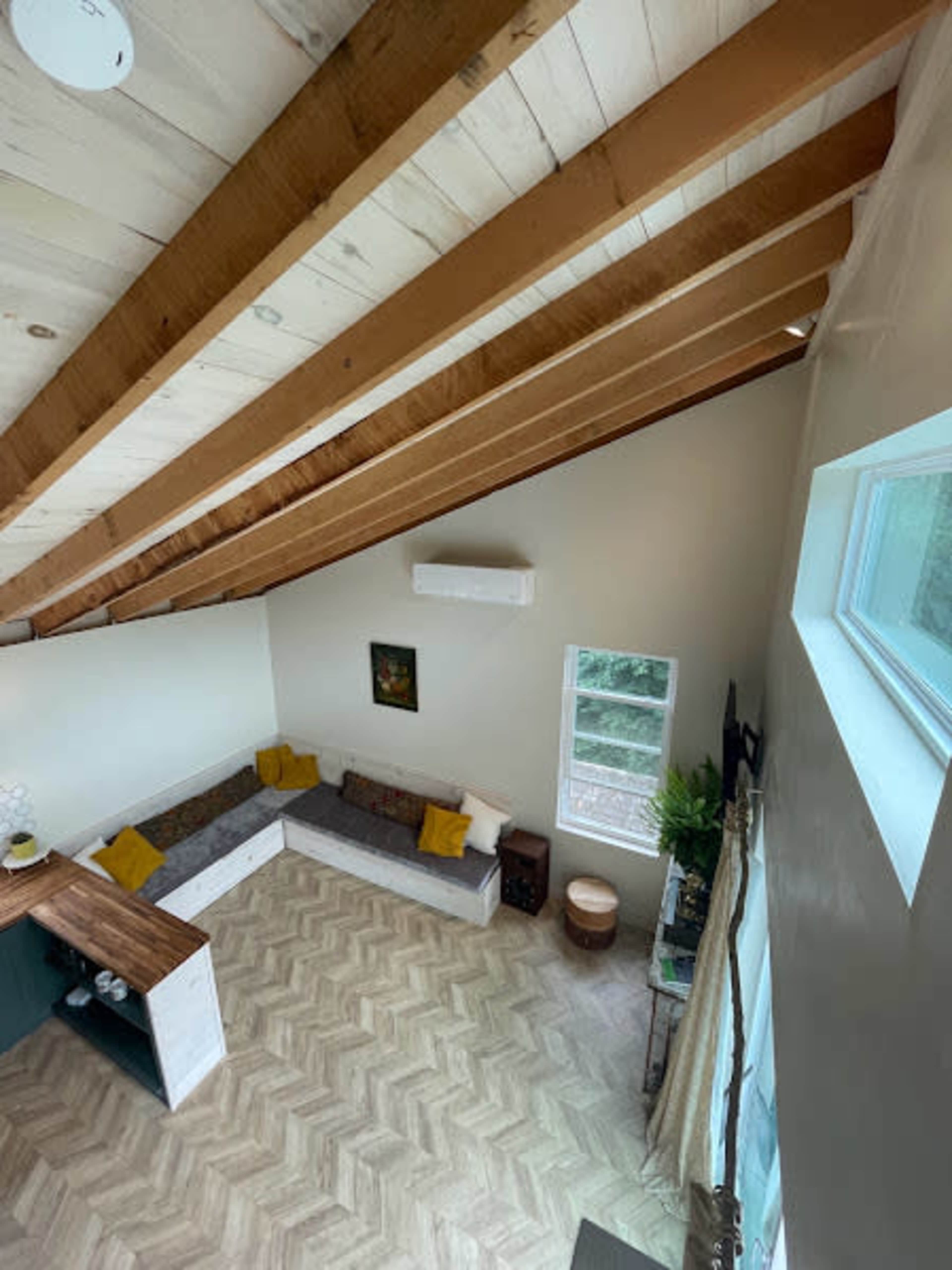 The image shows a spacious living room with a high, slanted ceiling and a corner sectional sofa accompanied by yellow cushions, a wooden coffee table, and a large window.