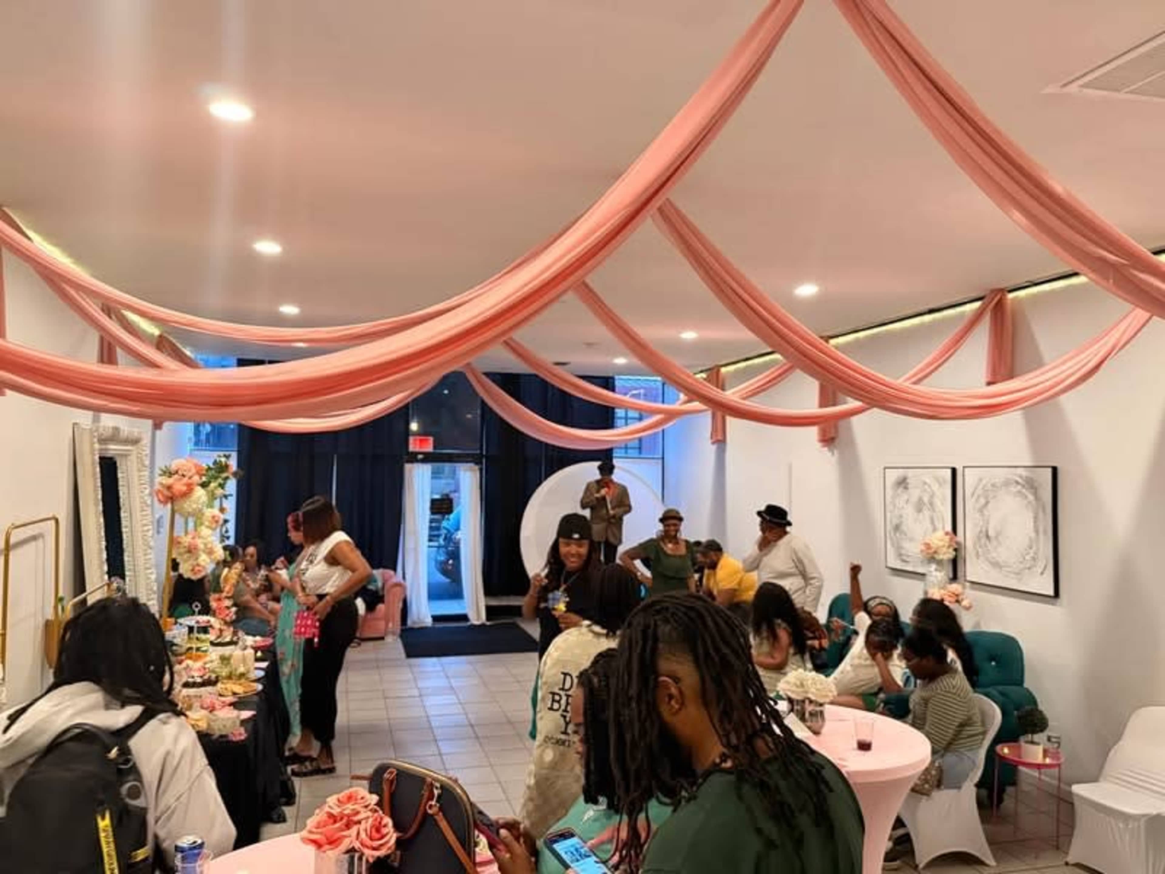 The scene depicts a lively event in a decorated indoor space, with guests mingling around tables adorned with snacks and floral arrangements under flowing fabric drapery.