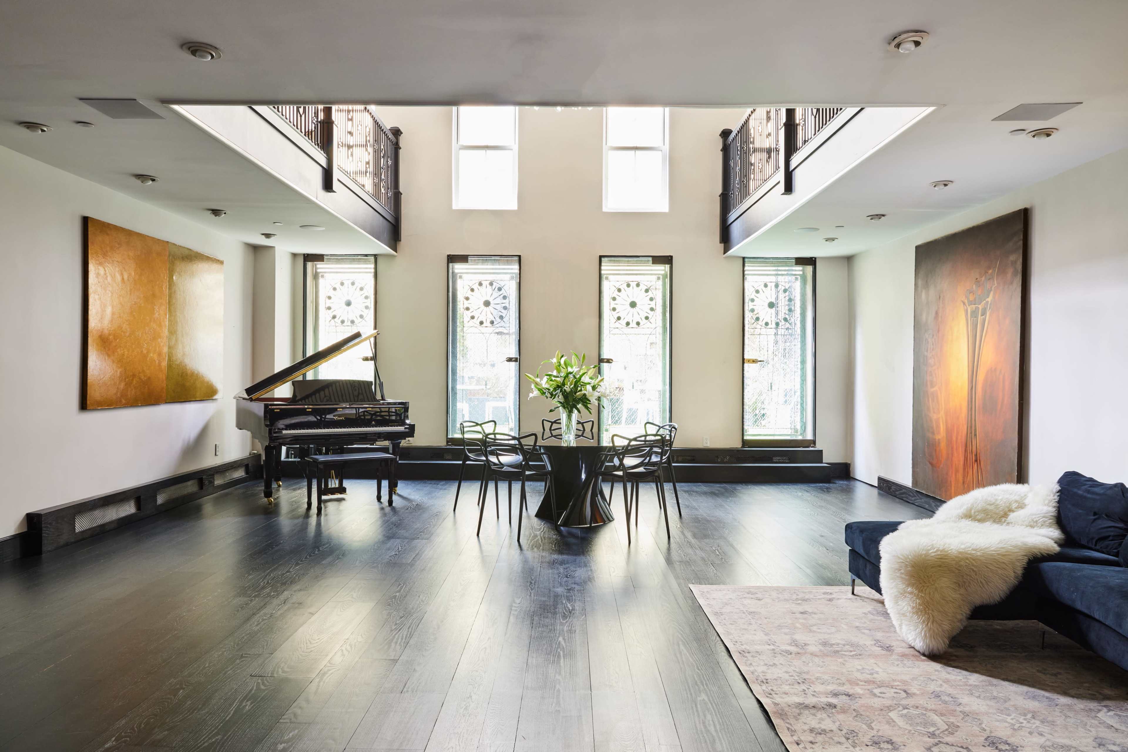 A spacious room features a grand piano, a round table with chairs, and large windows displaying ornate patterns, complemented by artwork on the walls.