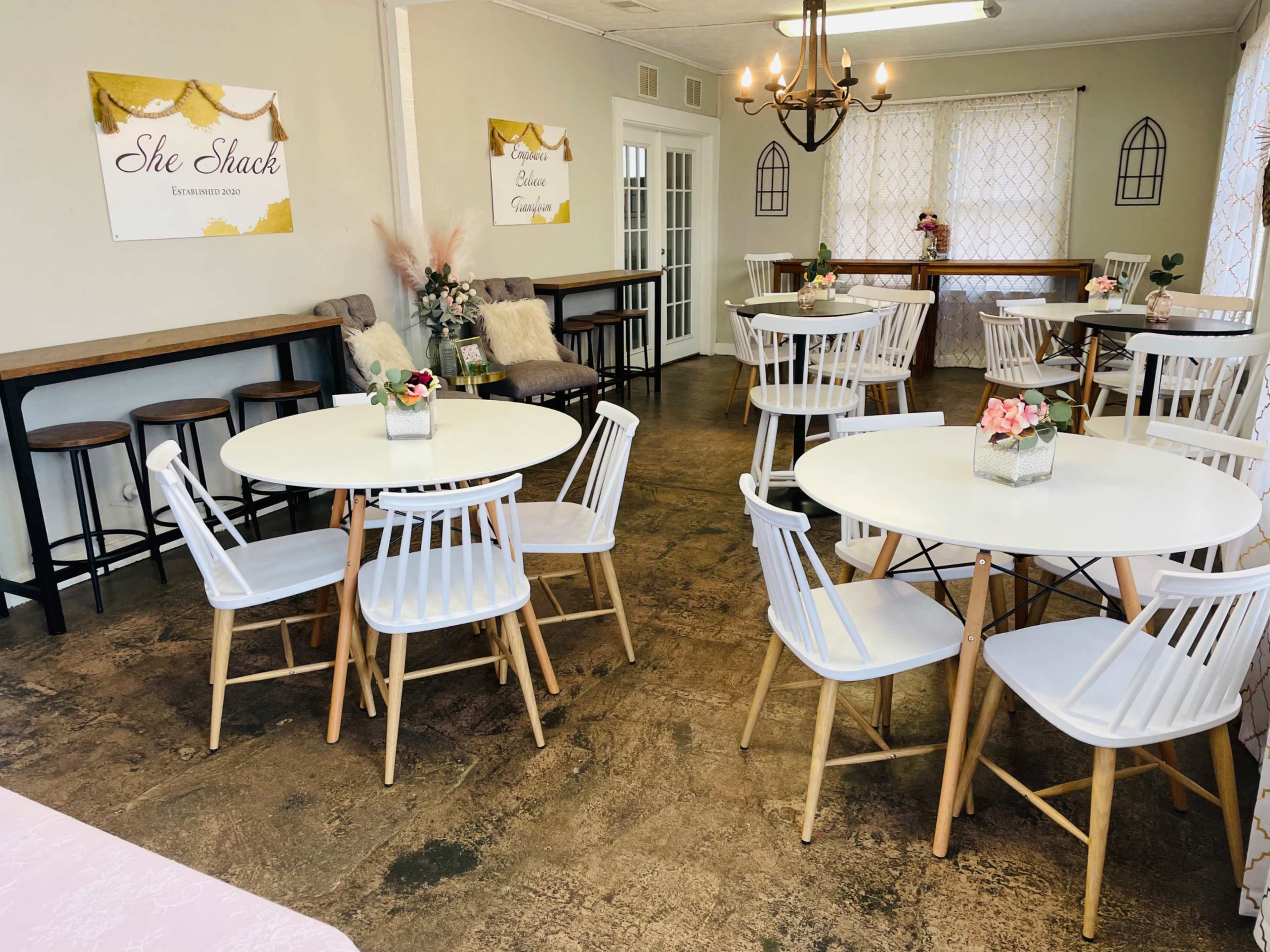 A cozy café interior with white tables and chairs arranged around the space, decorated with flower centerpieces and two posters on the walls.