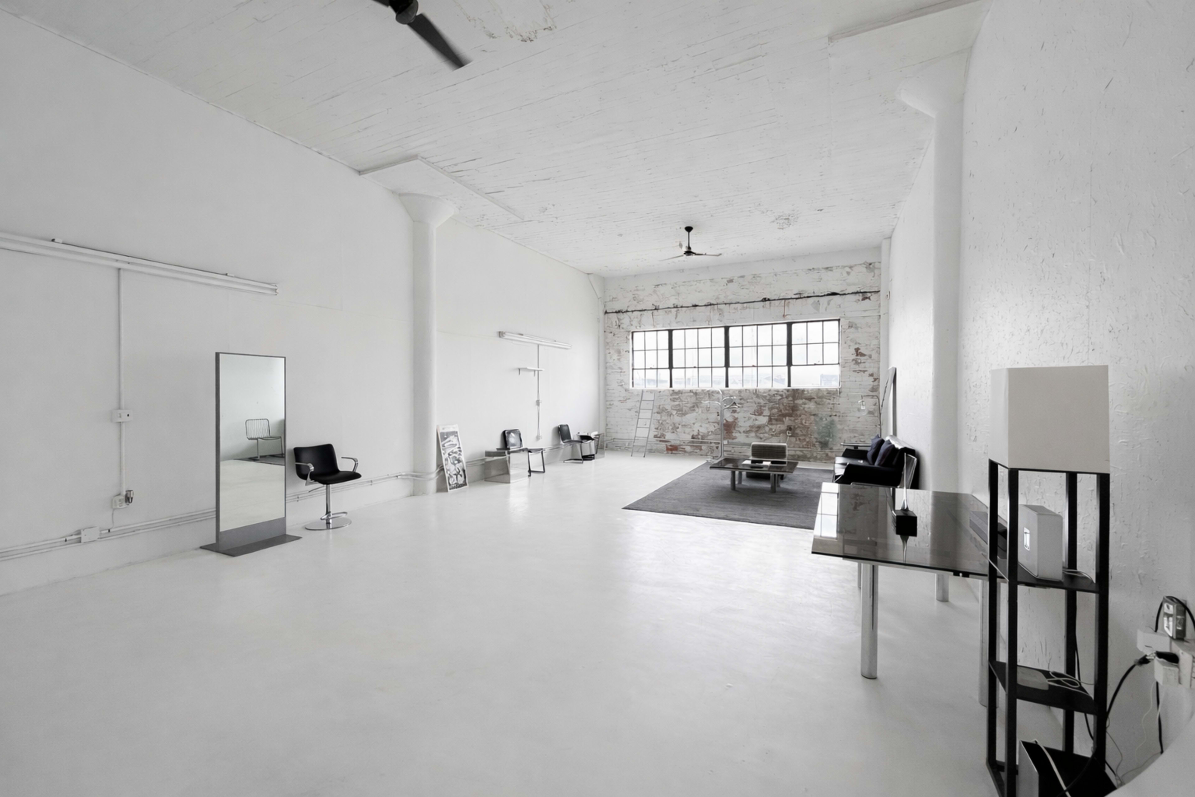 The image shows a spacious, minimalist interior with white walls and flooring, featuring a few pieces of modern furniture, including chairs and a table, and large windows allowing natural light.