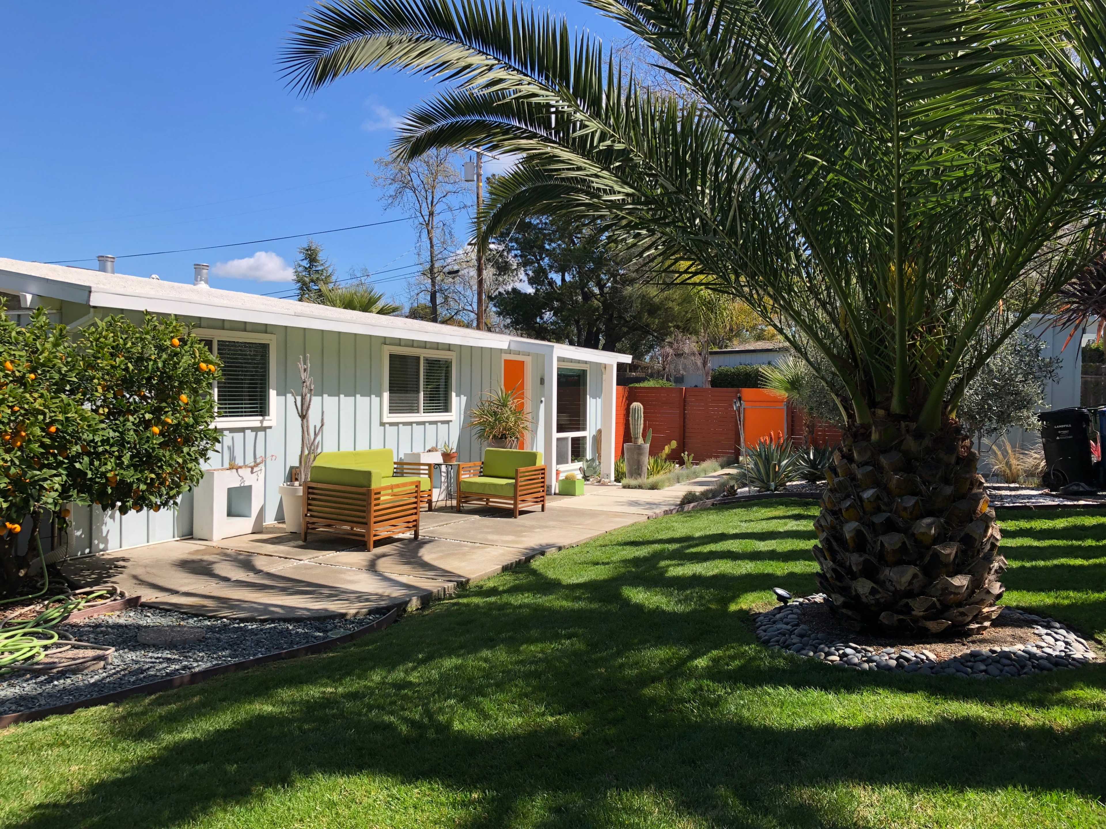 A small house with light blue siding features a patio with two wooden chairs, surrounded by palm trees and landscaped grass.