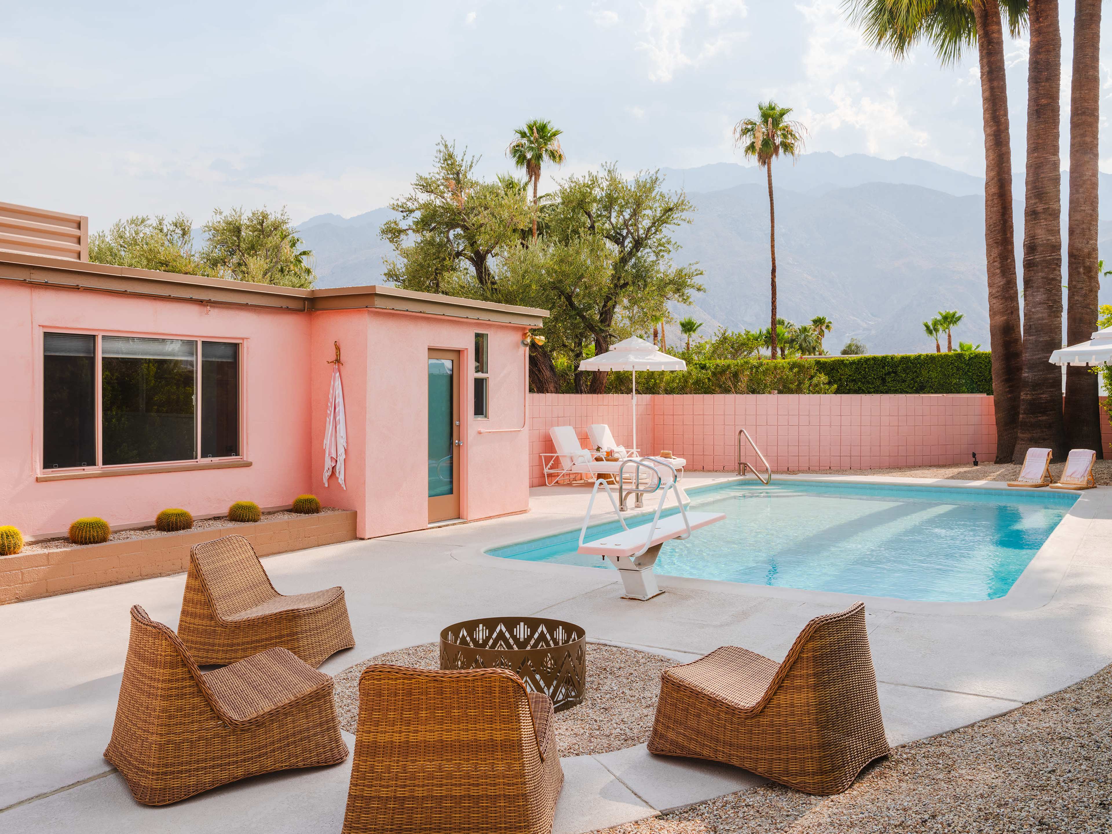 A pink house next to a swimming pool surrounded by lounge chairs and a fire pit, with palm trees and mountains in the background.