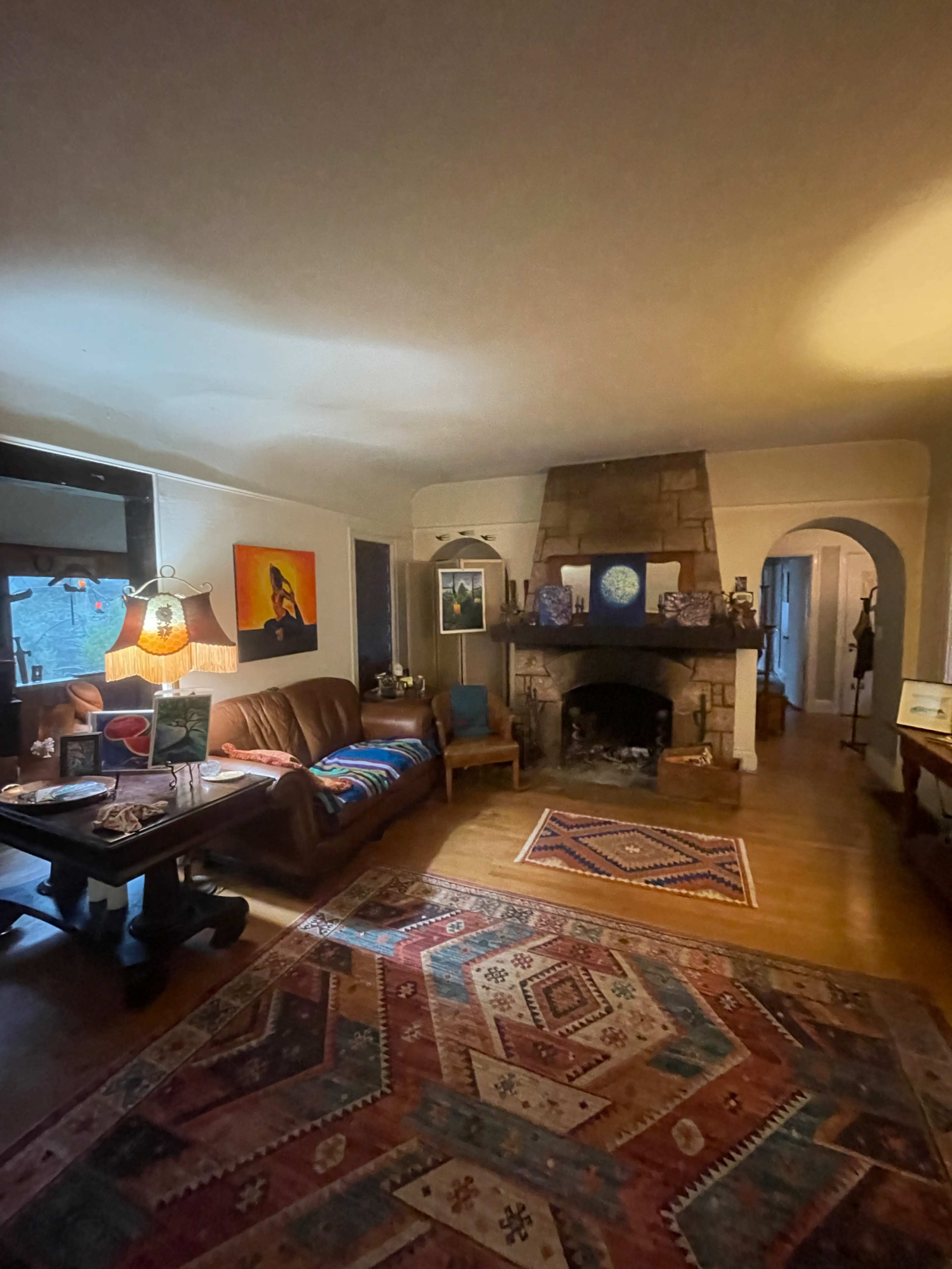 The image shows a cozy living room with a stone fireplace, a brown leather sofa, a colorful woven rug, and various artworks on the walls.