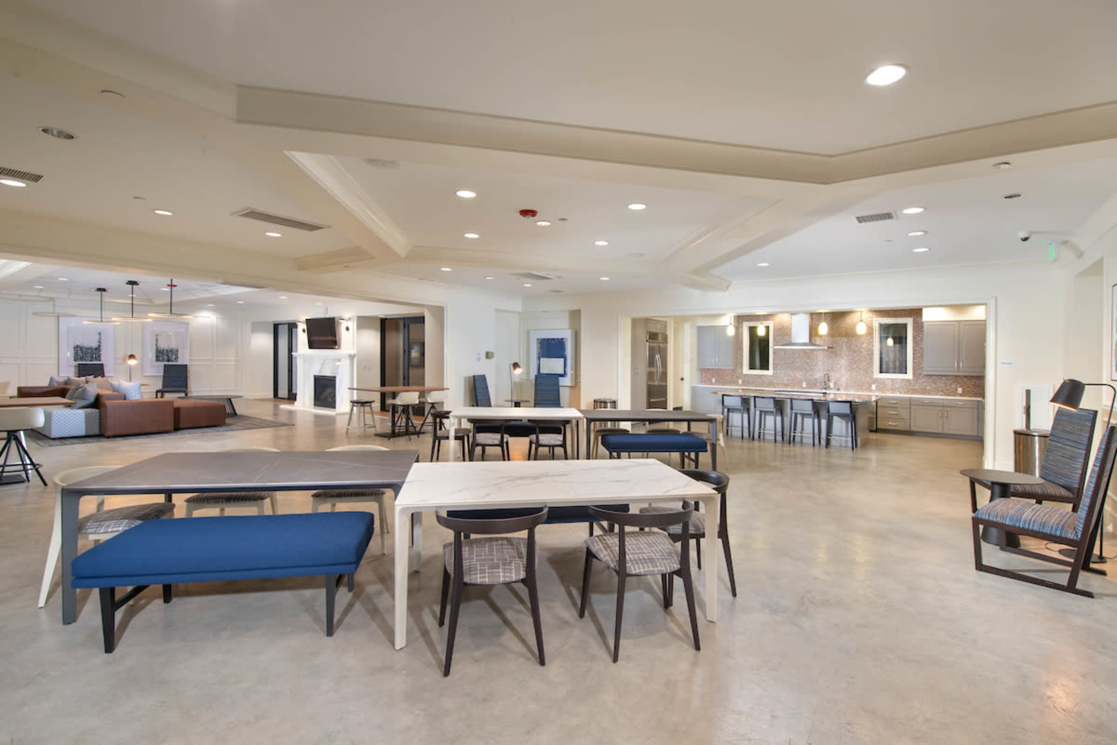 A spacious communal lounge area with various seating arrangements, including tables and chairs, and a kitchen counter in the background.
