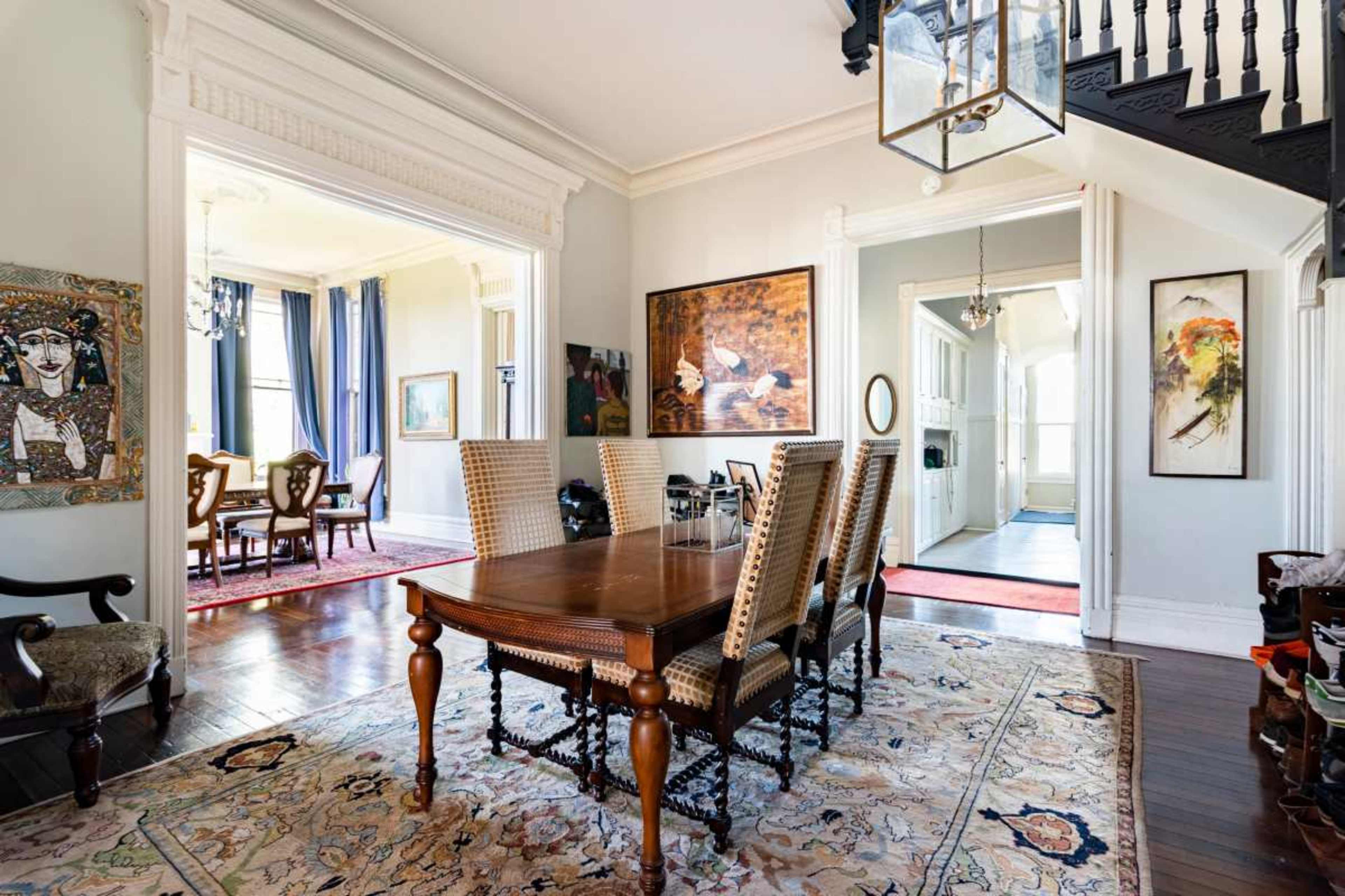 The image shows a spacious dining area with a wooden table surrounded by high-backed chairs, decorated with artwork and a large patterned rug.