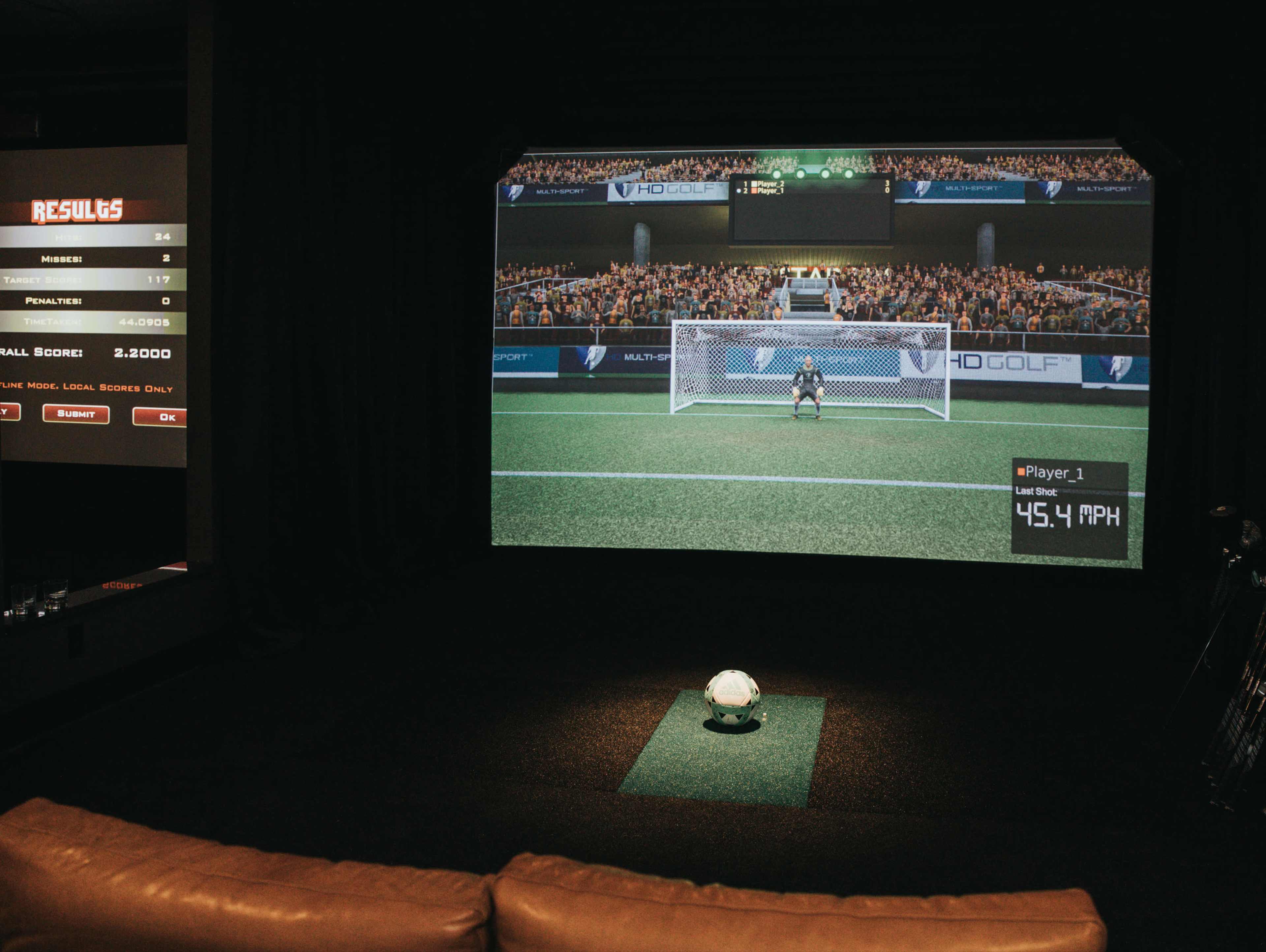 A large projection screen displays a soccer game simulation, with a ball placed on the turf in front of it, while a scoreboard lists player results on the side.