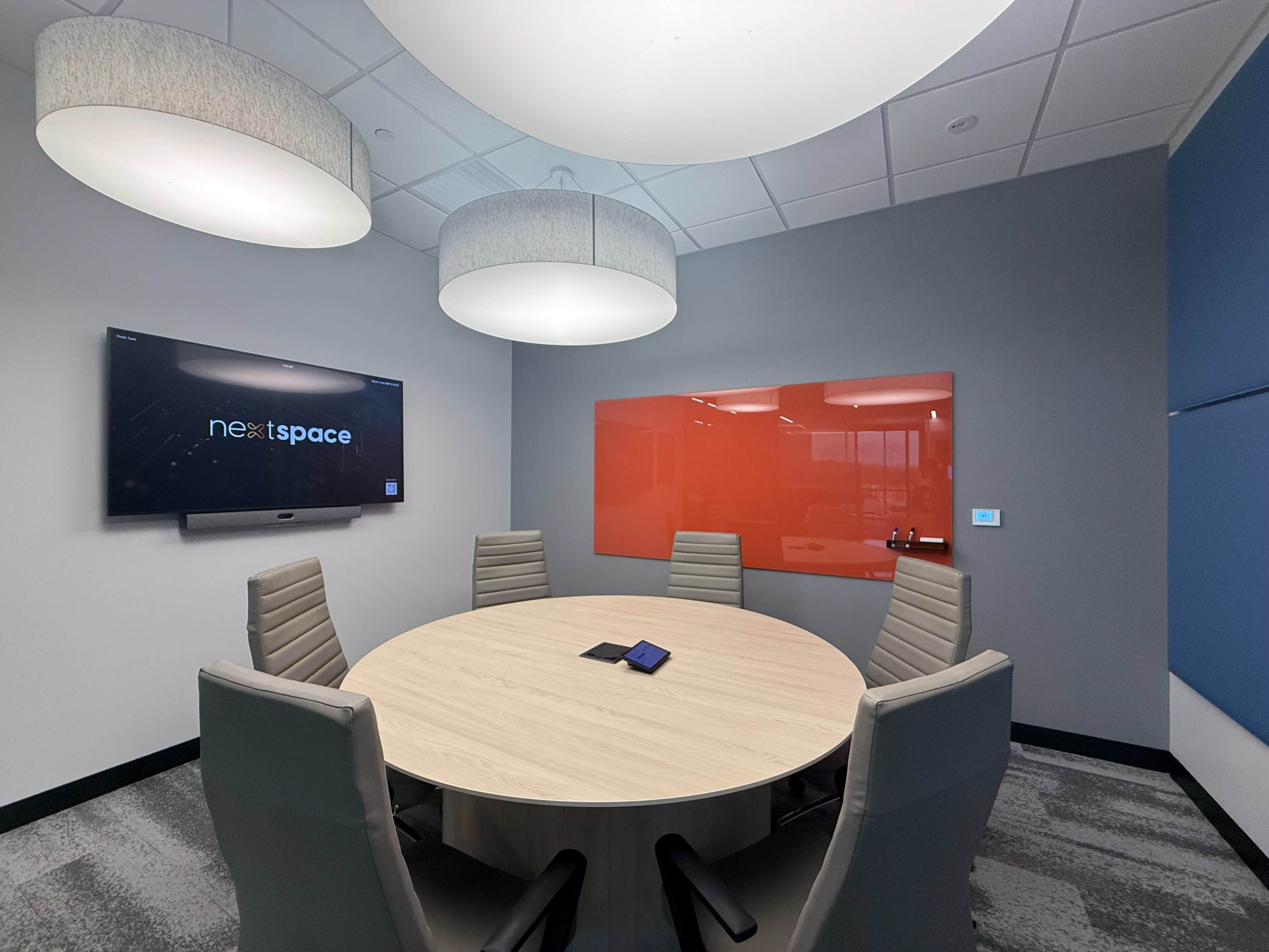 The image shows a modern conference room with a circular table, six gray chairs, a large screen on the wall, and a red glass board.