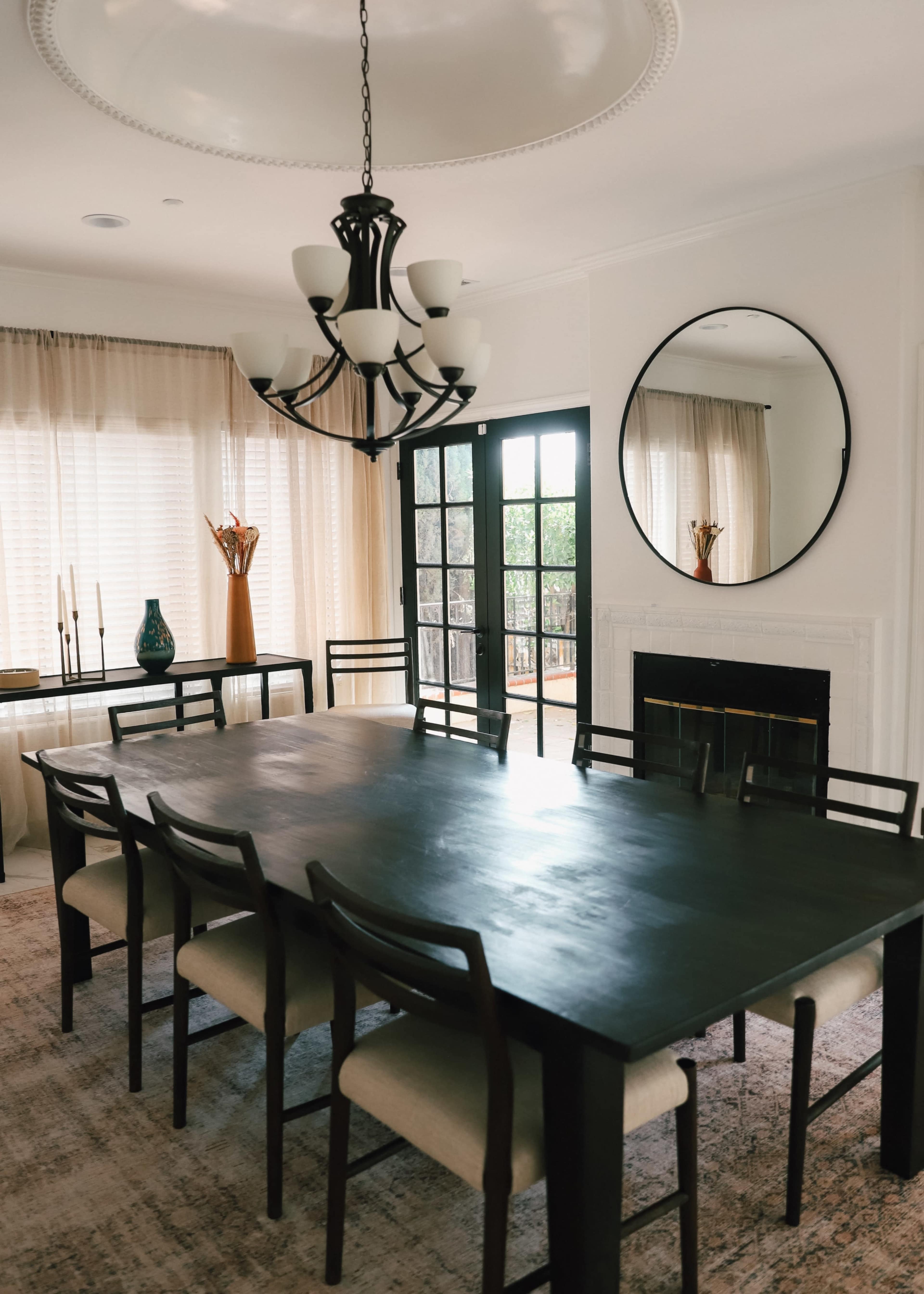 A long dark wooden dining table with eight chairs is situated in a well-lit room featuring large windows and a round mirror on the wall.