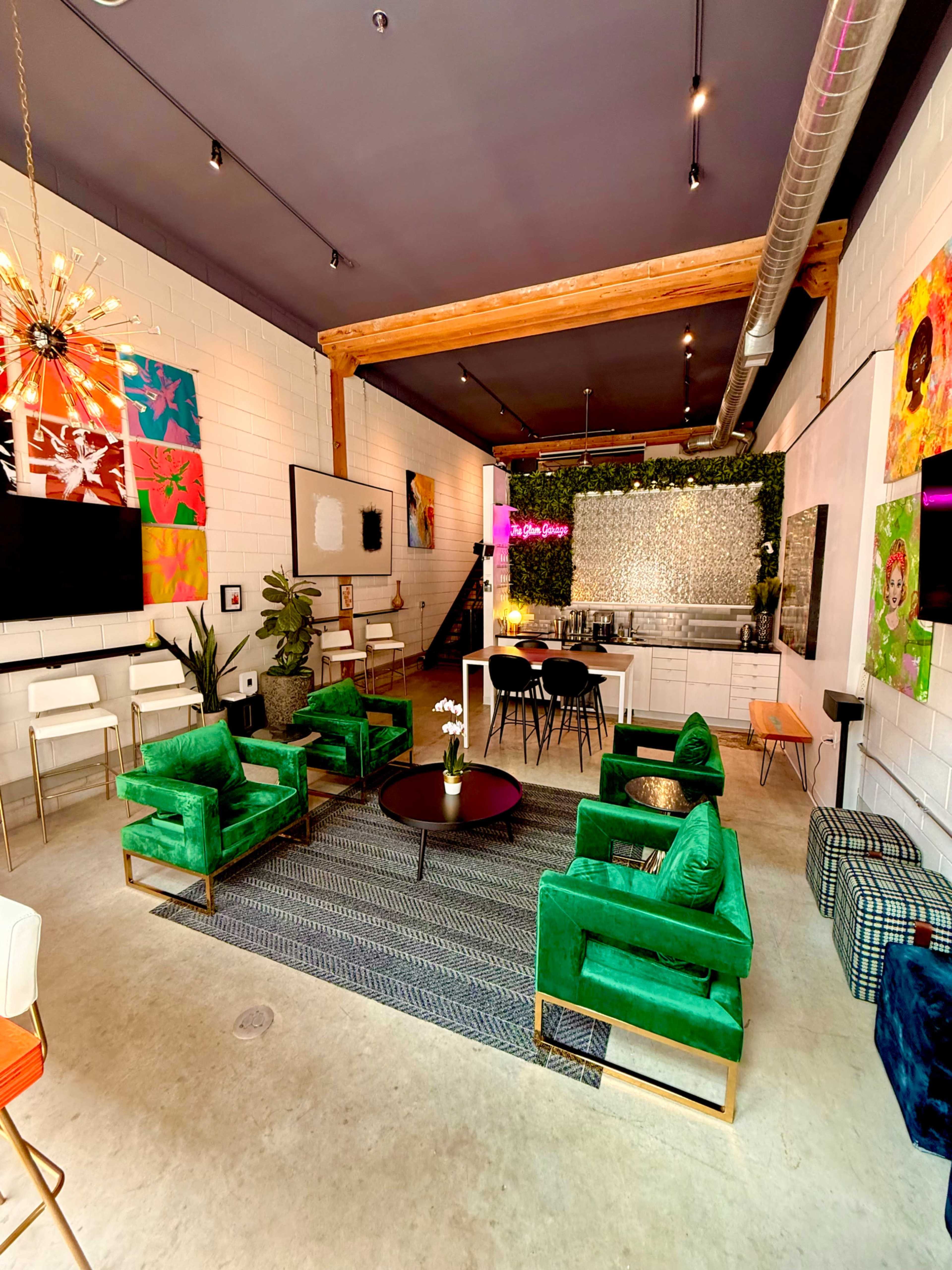 The image shows a modern indoor space featuring green upholstered chairs, a dining area, a kitchen with a bar, and vibrant artwork on the walls.