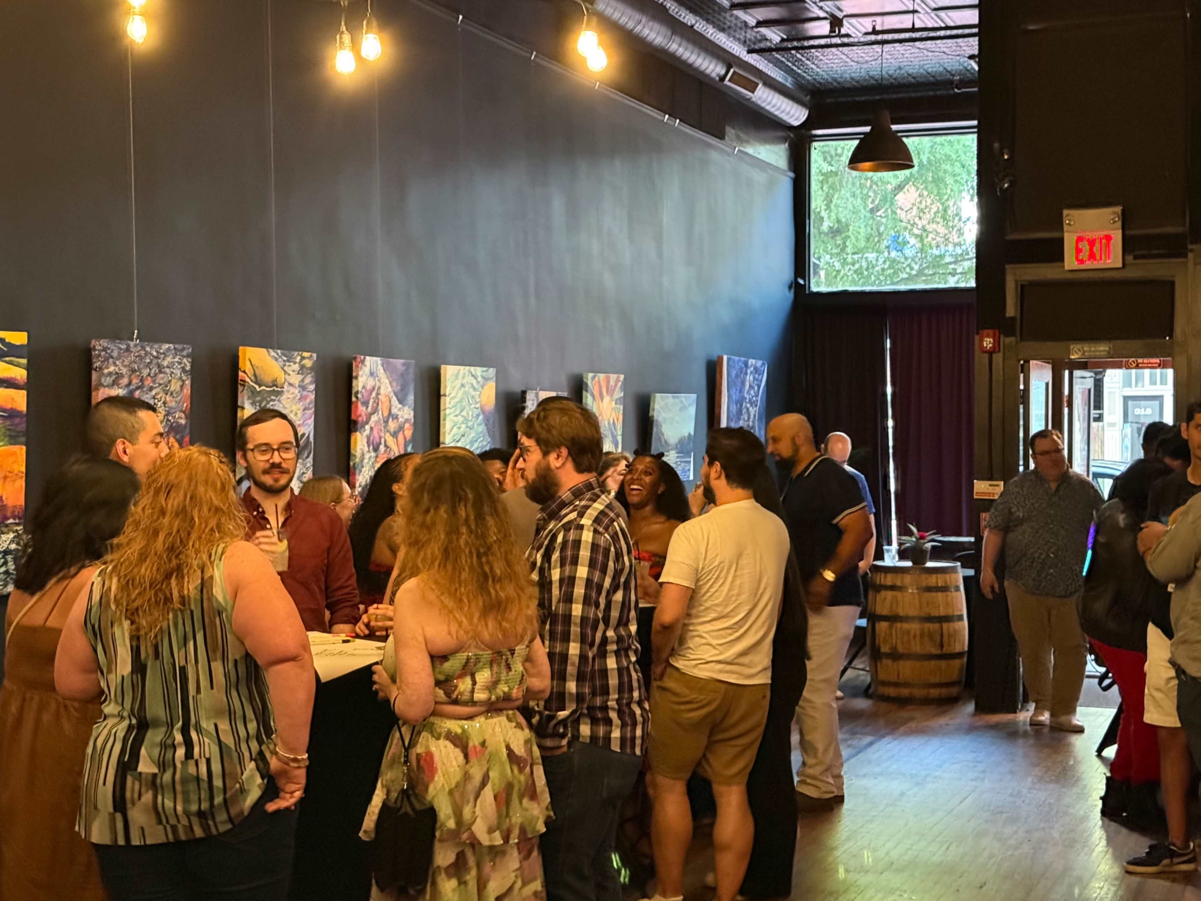 A group of people is gathered in a dimly lit art gallery, engaging in conversation near a display of paintings on the wall.