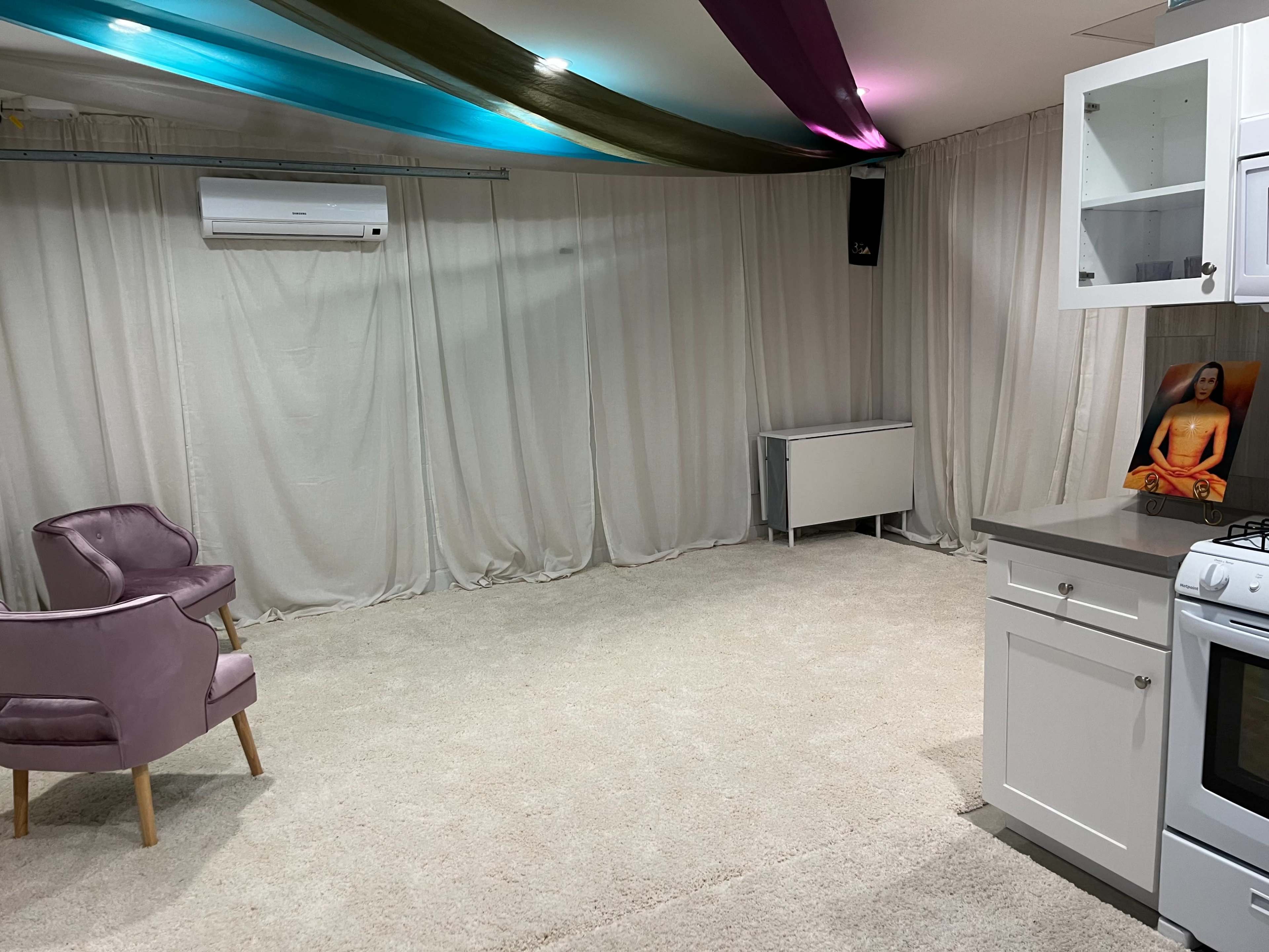 The image shows a spacious room with beige carpeting, a small kitchen area featuring white cabinets and a stove, two pink chairs, and draped curtains in the background.