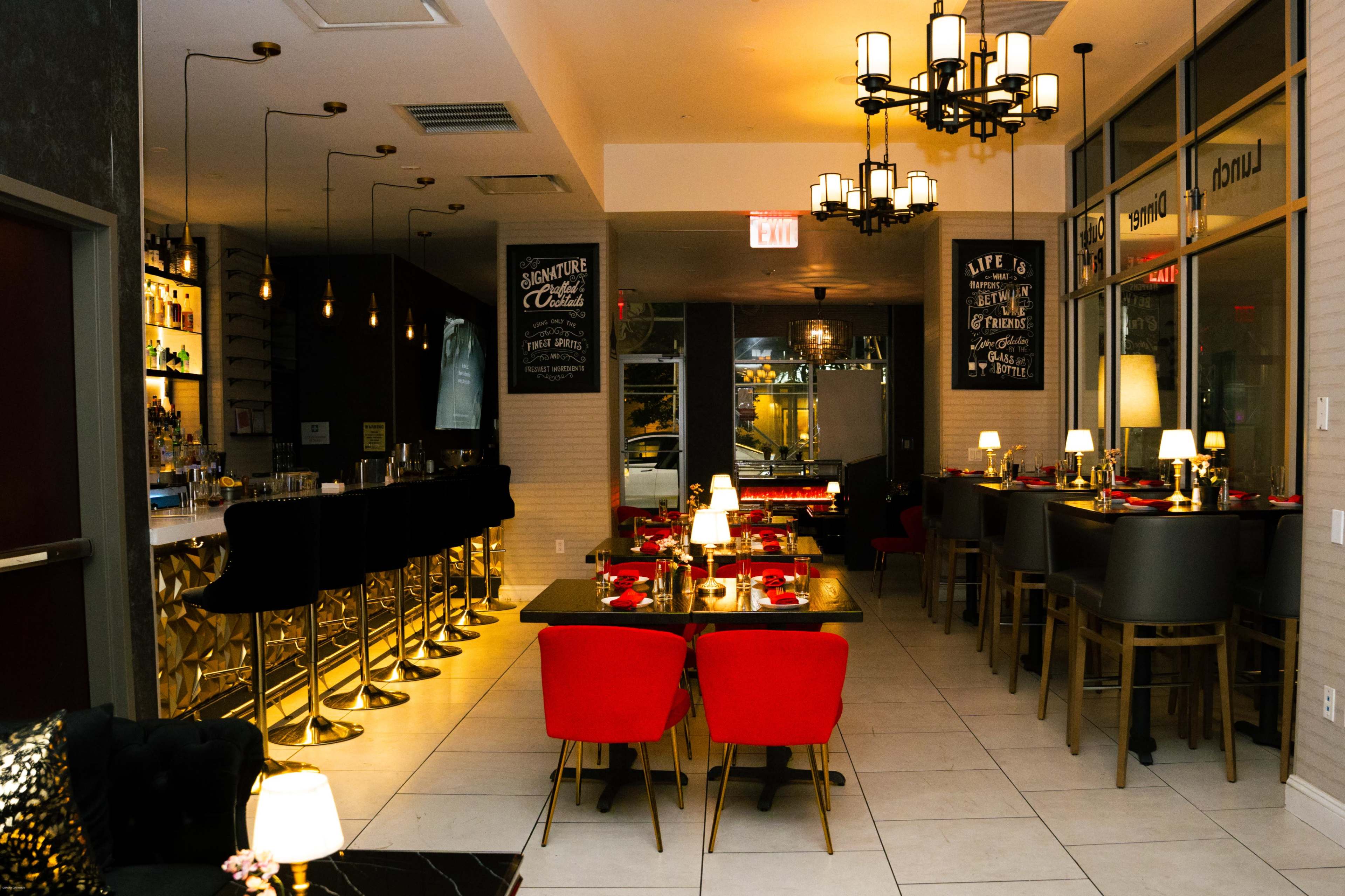 The image shows a stylish restaurant interior featuring a bar area with high stools, elegantly set tables with red and black accents, and ambient lighting.