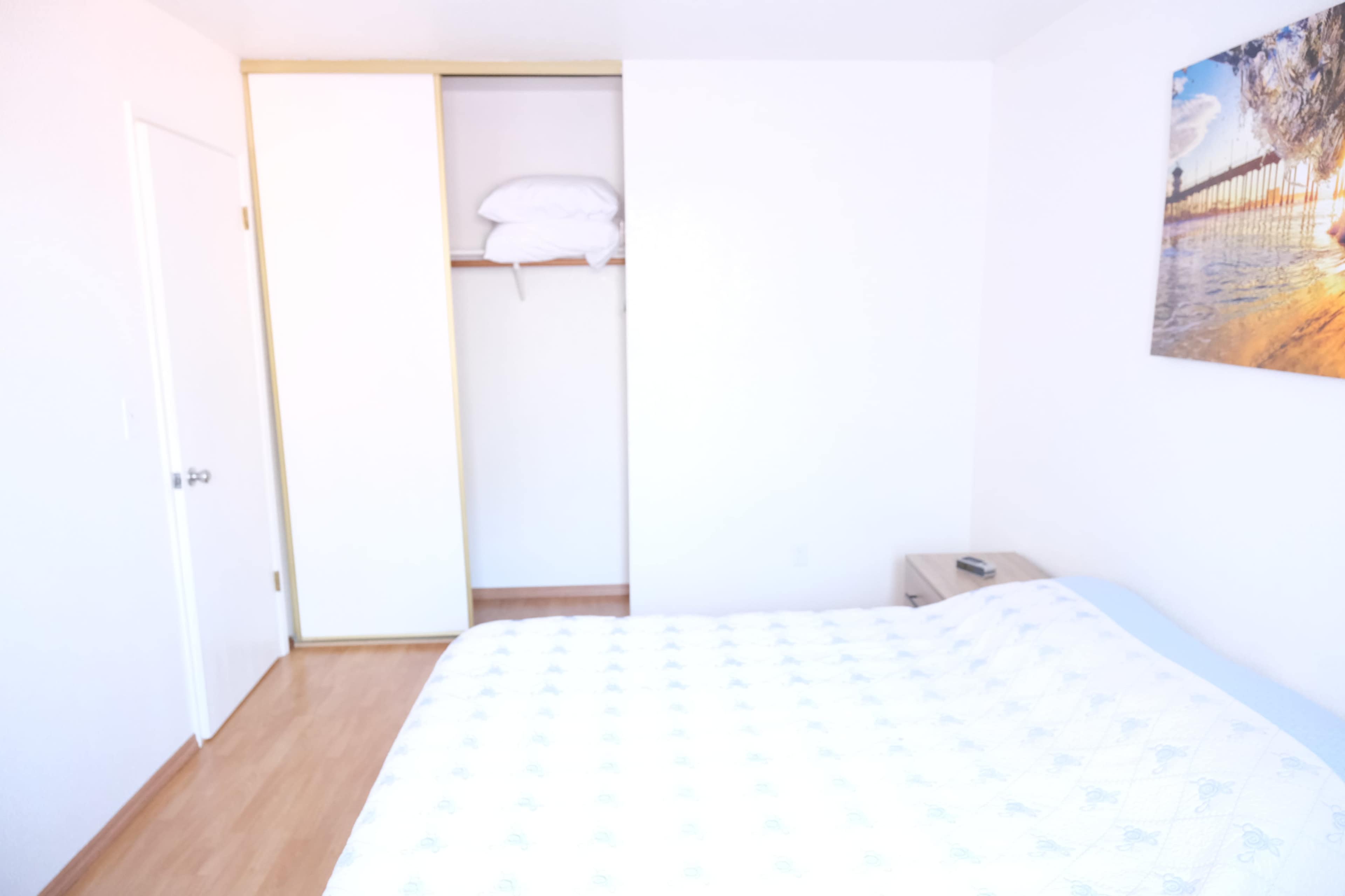 The image shows a bedroom with a bed, a nightstand, and an open closet displaying stacked pillows.