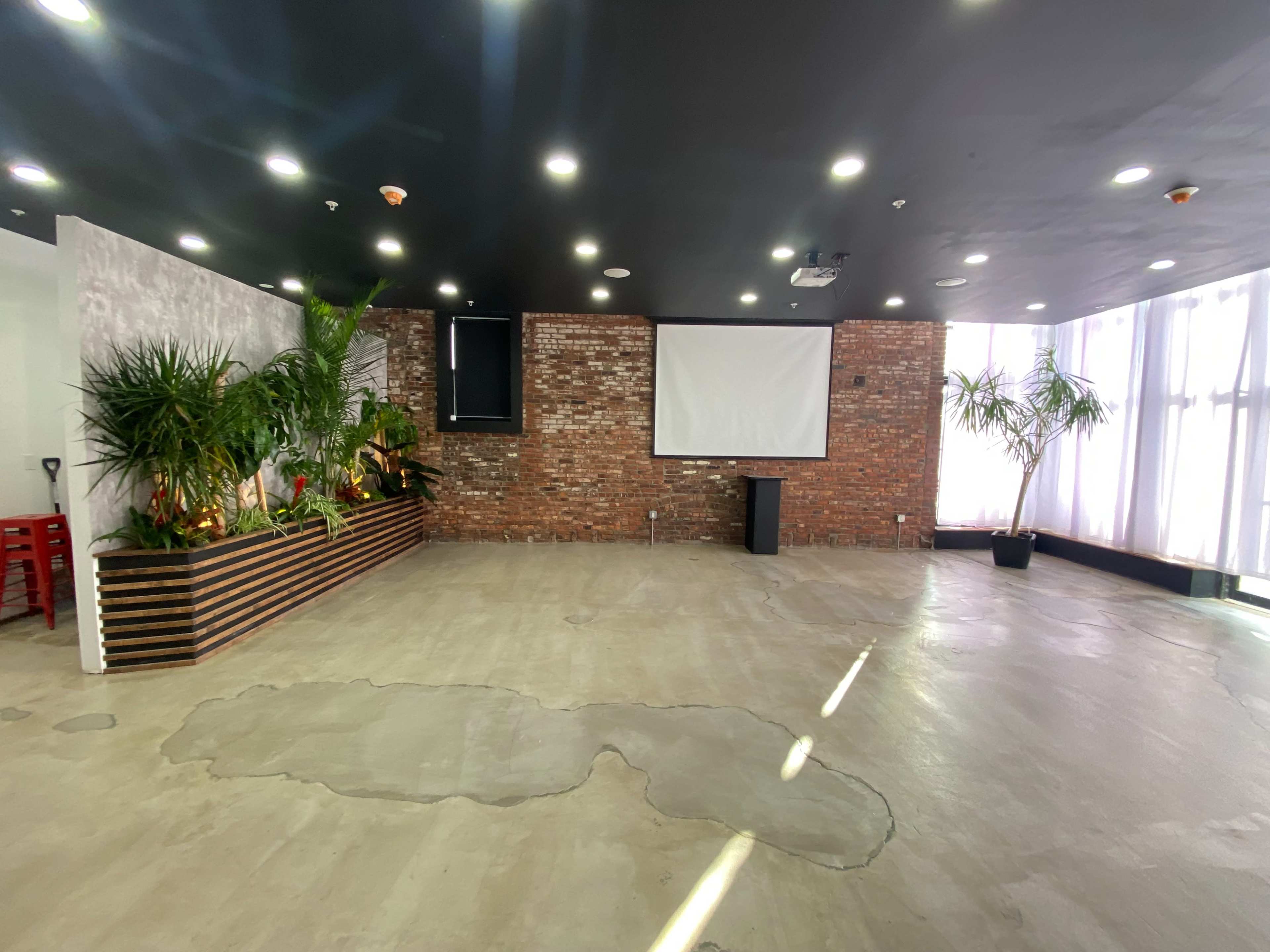 The image shows a spacious, empty room with a large projection screen, brick walls, and tropical plants in wooden planters.