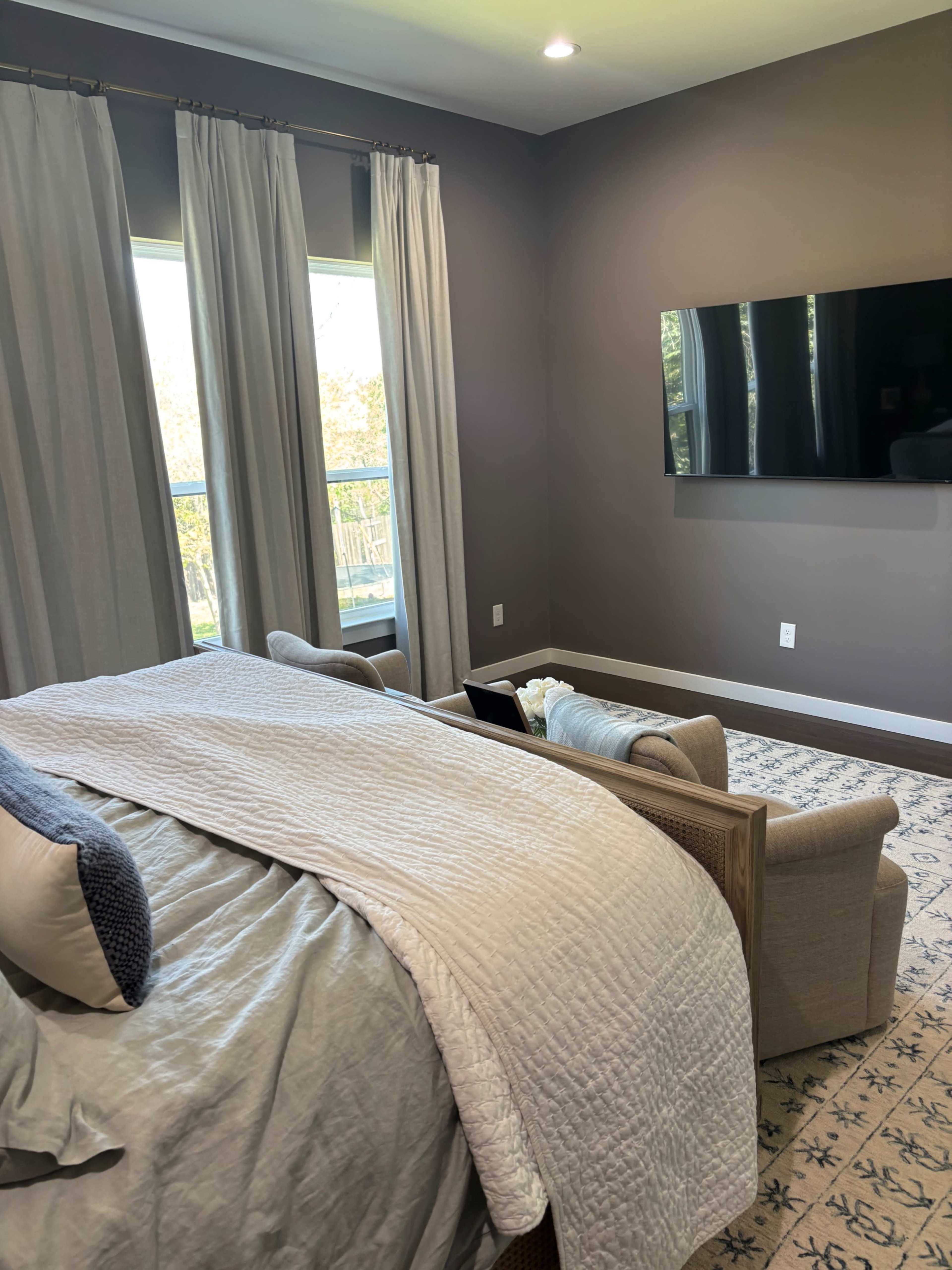 The image shows a bedroom with a large bed covered in a light quilt, two chairs facing a wall-mounted television, and large windows with sheer curtains.
