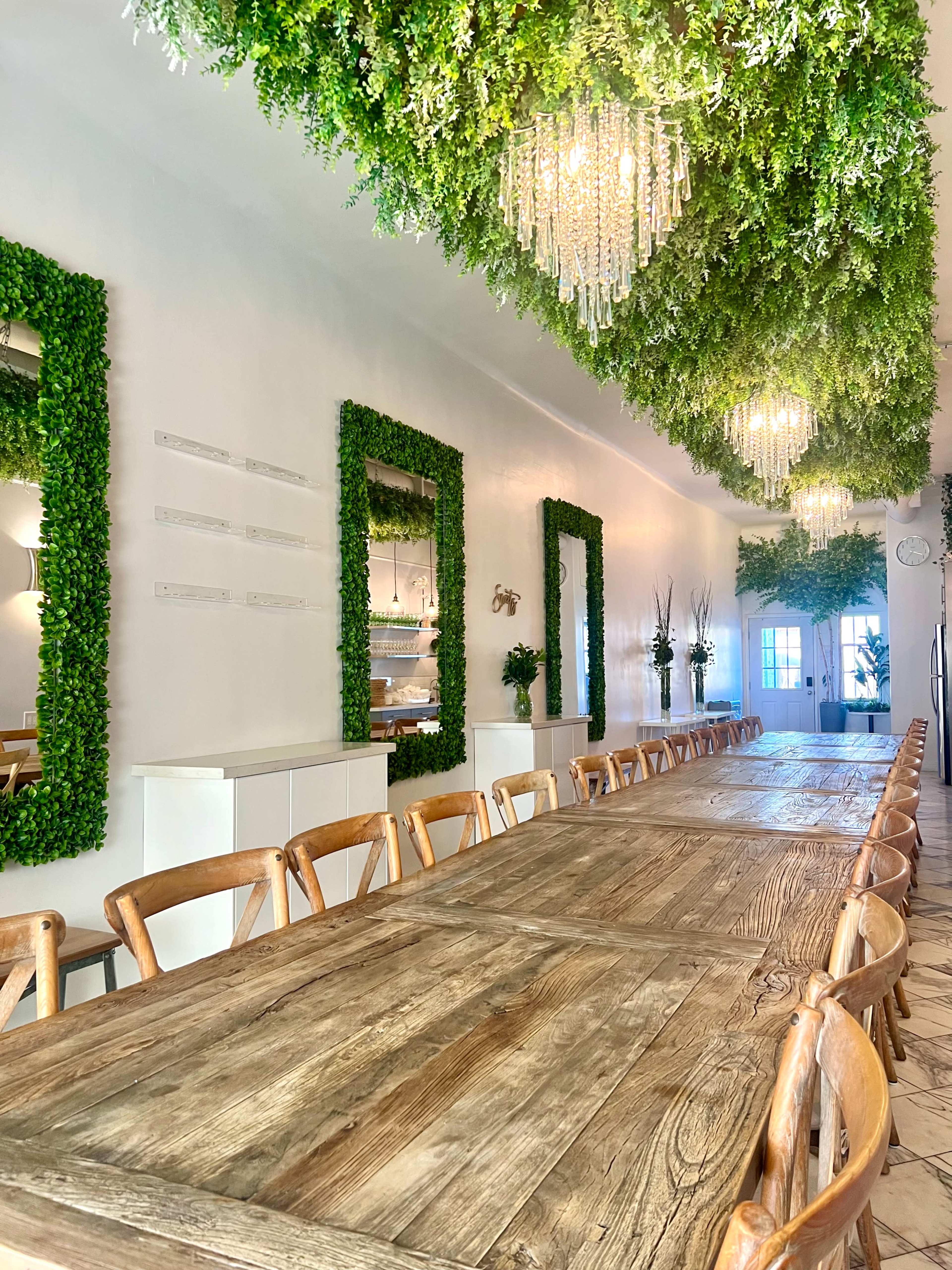 A long wooden table with wooden chairs is centered in a bright room, which features green wall decor and hanging plants, along with elegant chandeliers.