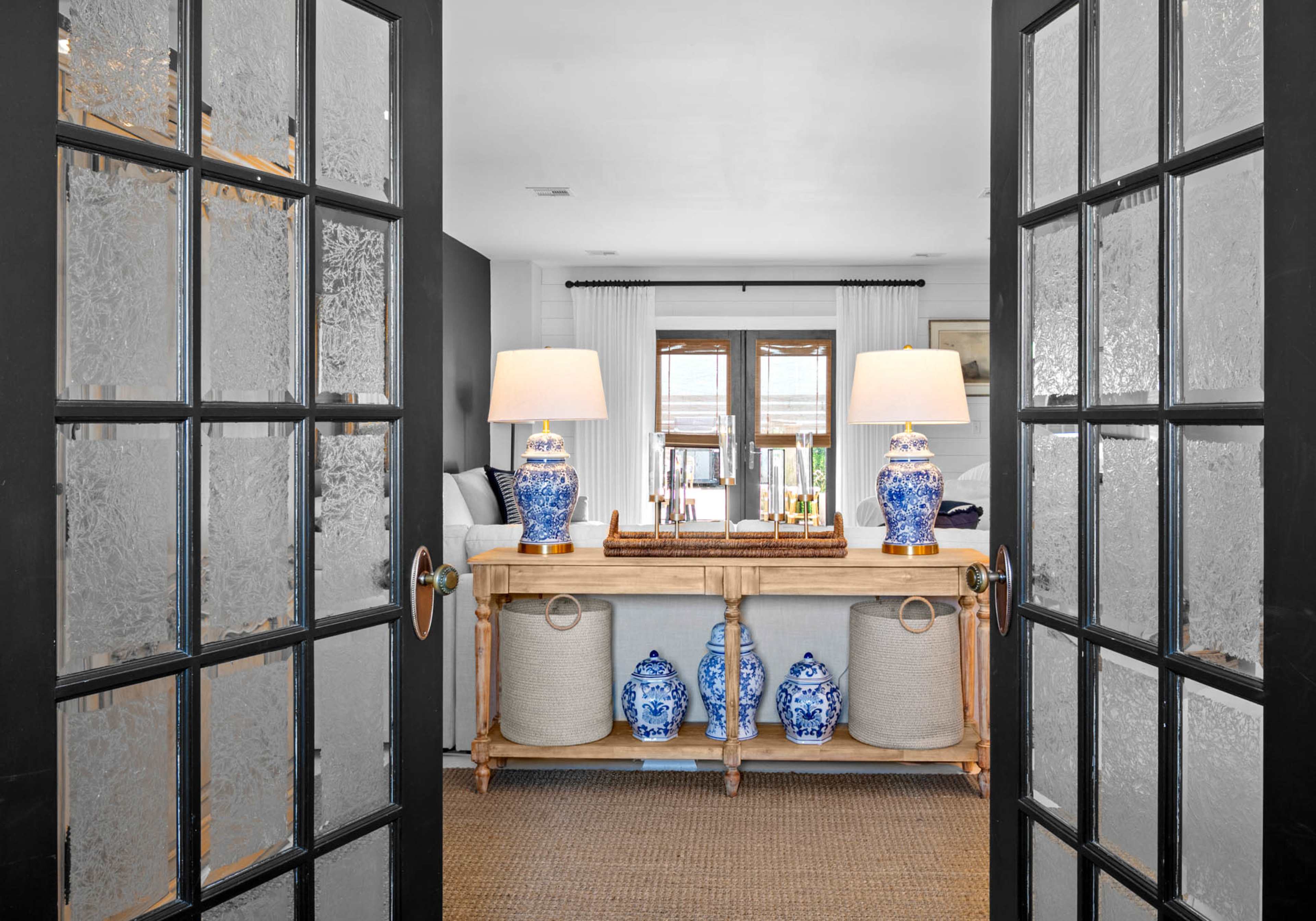 The image shows an entrance through black-framed glass doors into a bright living space featuring a console table with blue and white decor, two lamps, and decorative containers.