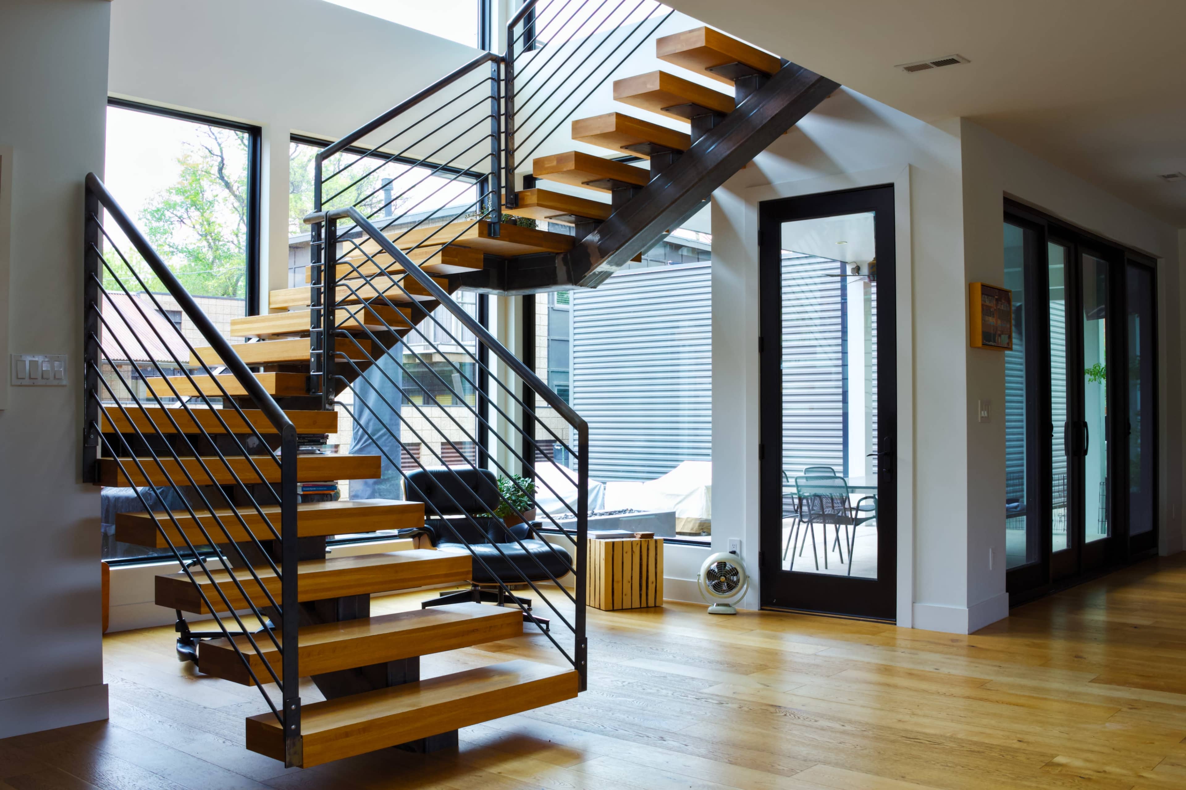 A modern staircase made of wood and metal is positioned in a well-lit interior space with large windows and an open layout.