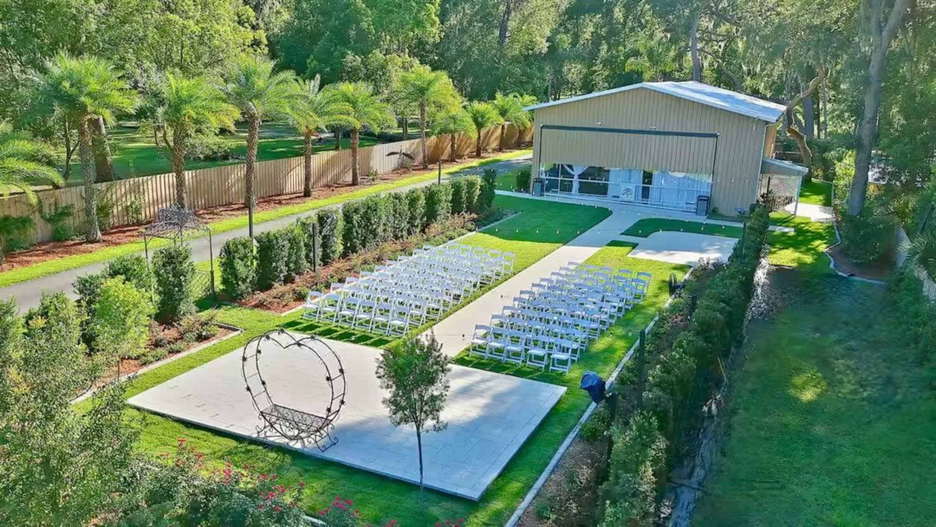 The image shows an outdoor wedding venue with rows of white chairs arranged on a paved area in front of a barn surrounded by green landscaping and palm trees.