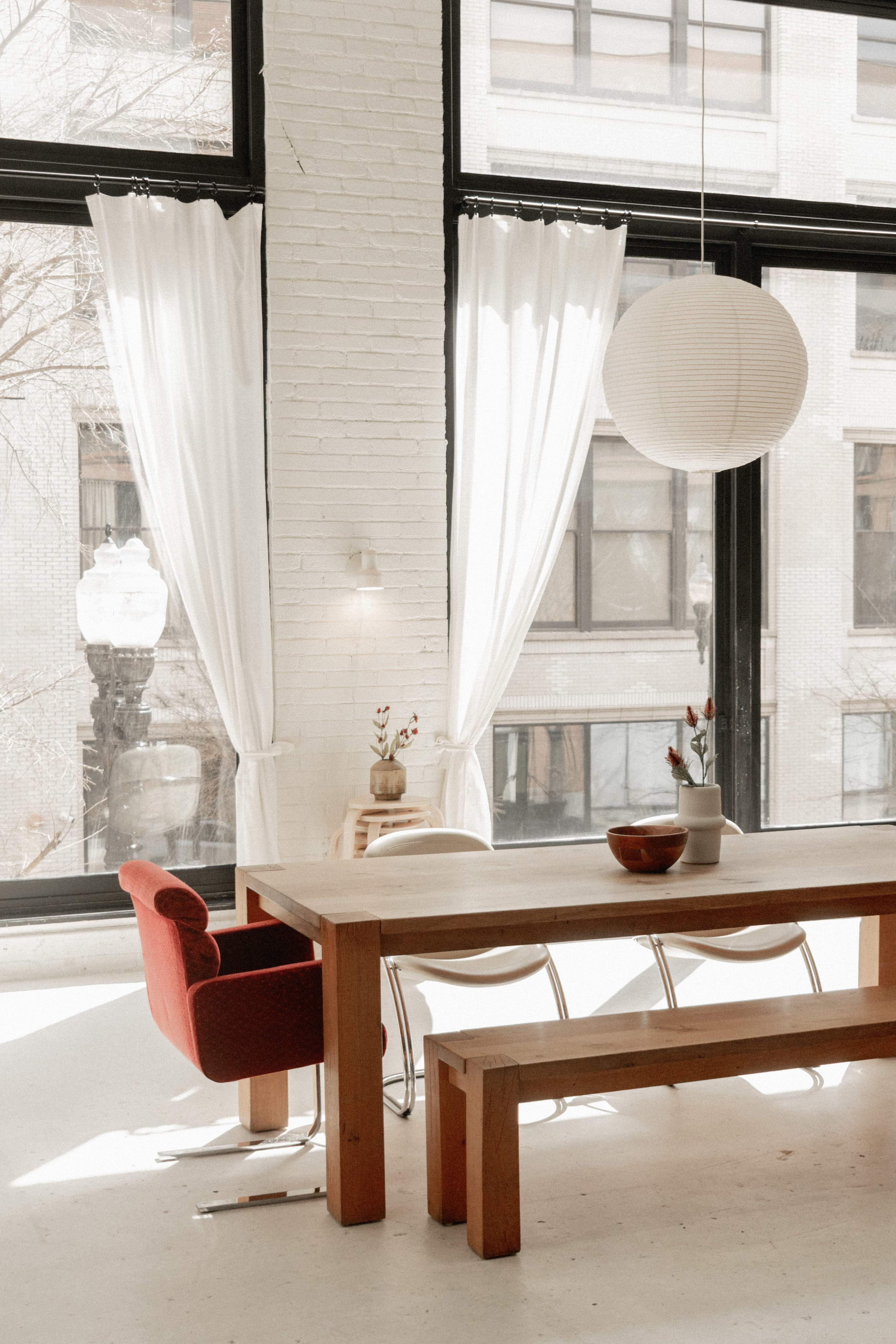 A spacious dining area features a long wooden table with a red chair, large windows dressed with white curtains, and a paper lantern overhead.