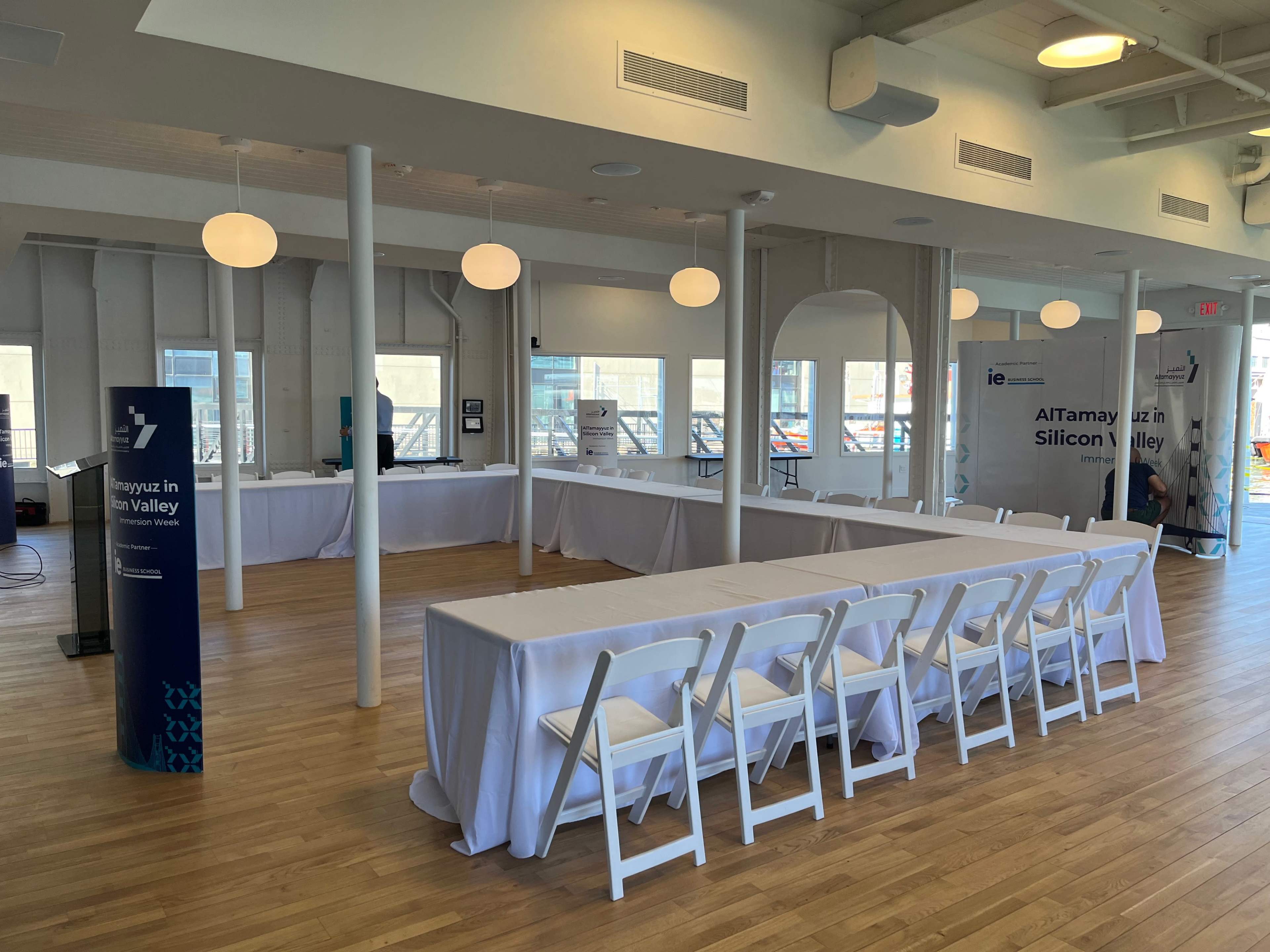 A spacious, well-lit event room with white tables covered in linens and a few promotional banners positioned along the walls.