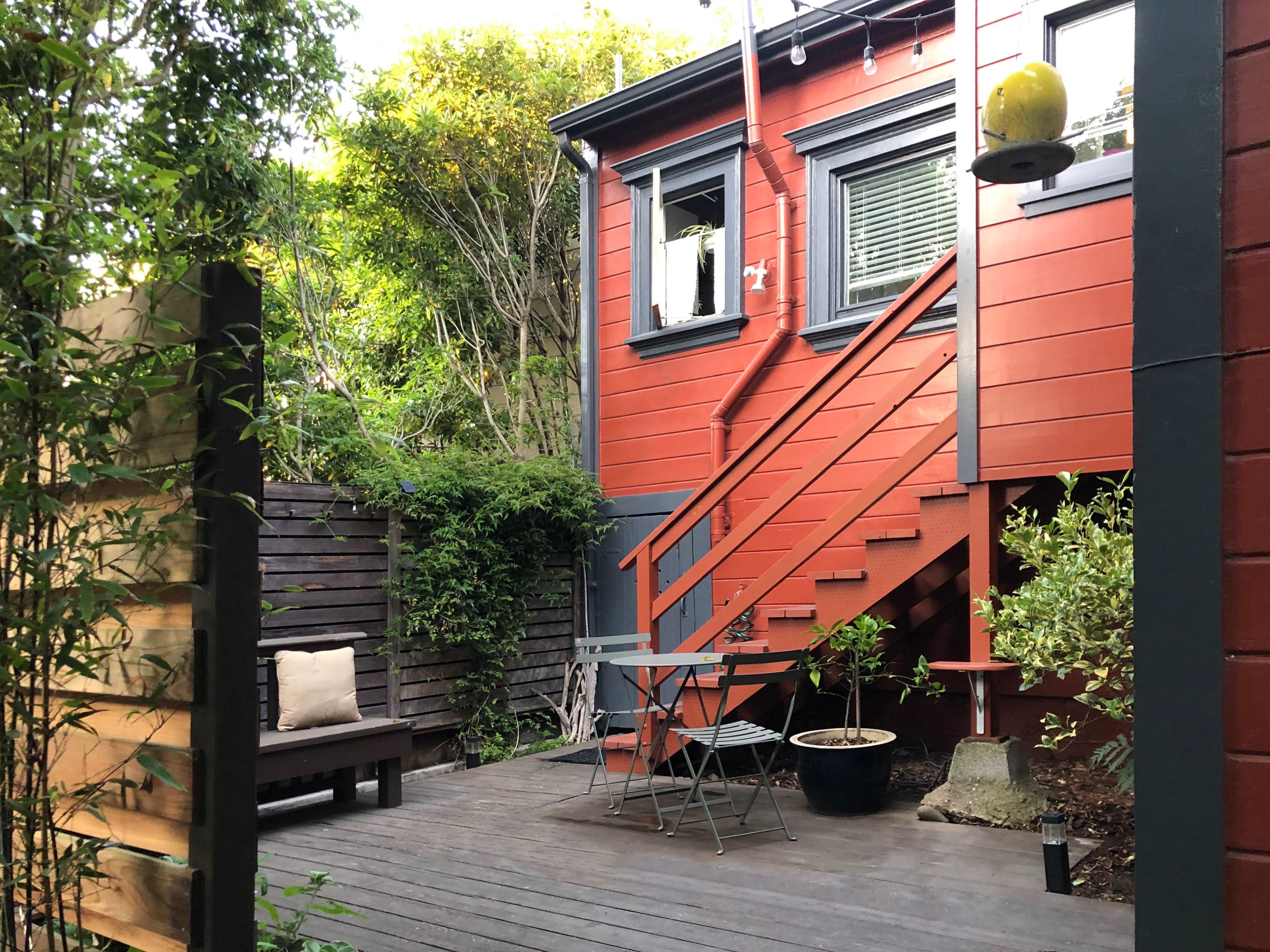 A small outdoor area with a wooden deck, a seating arrangement, and a red house with a staircase and green foliage surrounding it.
