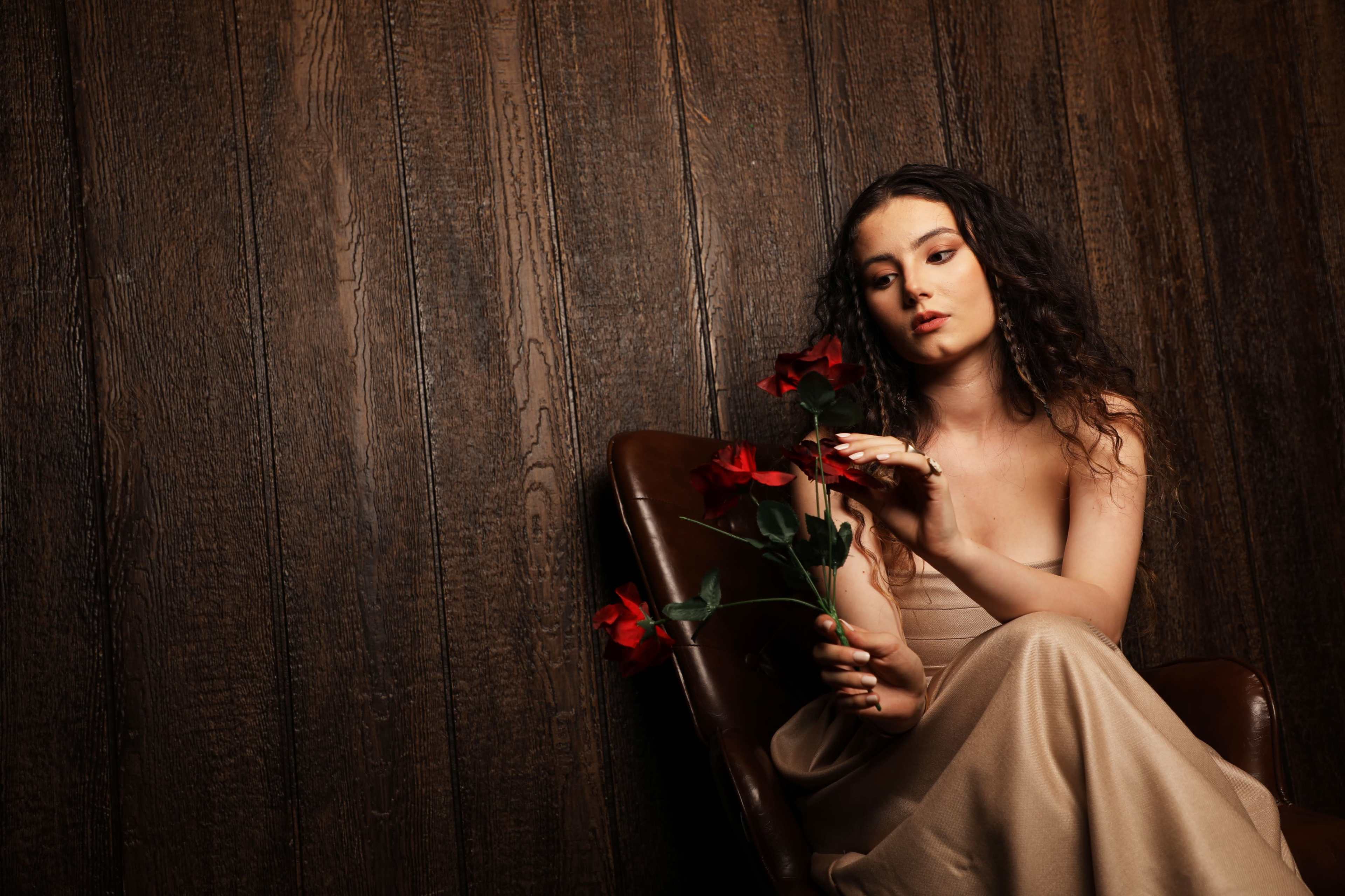 A woman in a strapless dress sits in a brown chair, holding red roses against a wooden backdrop.