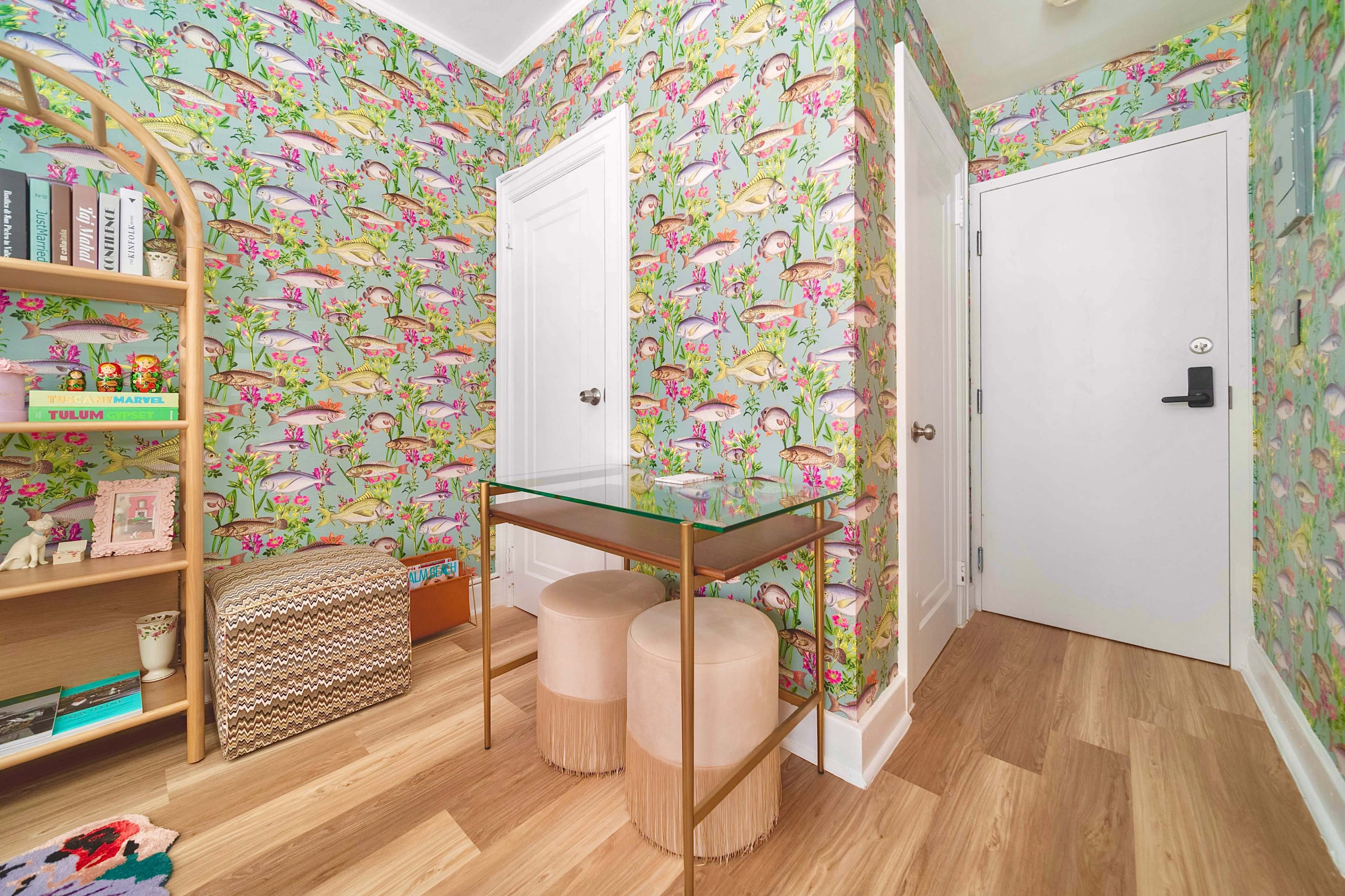 A corner of a room featuring vibrant floral wallpaper, a small glass table, two round stools, and a bookshelf against one wall.