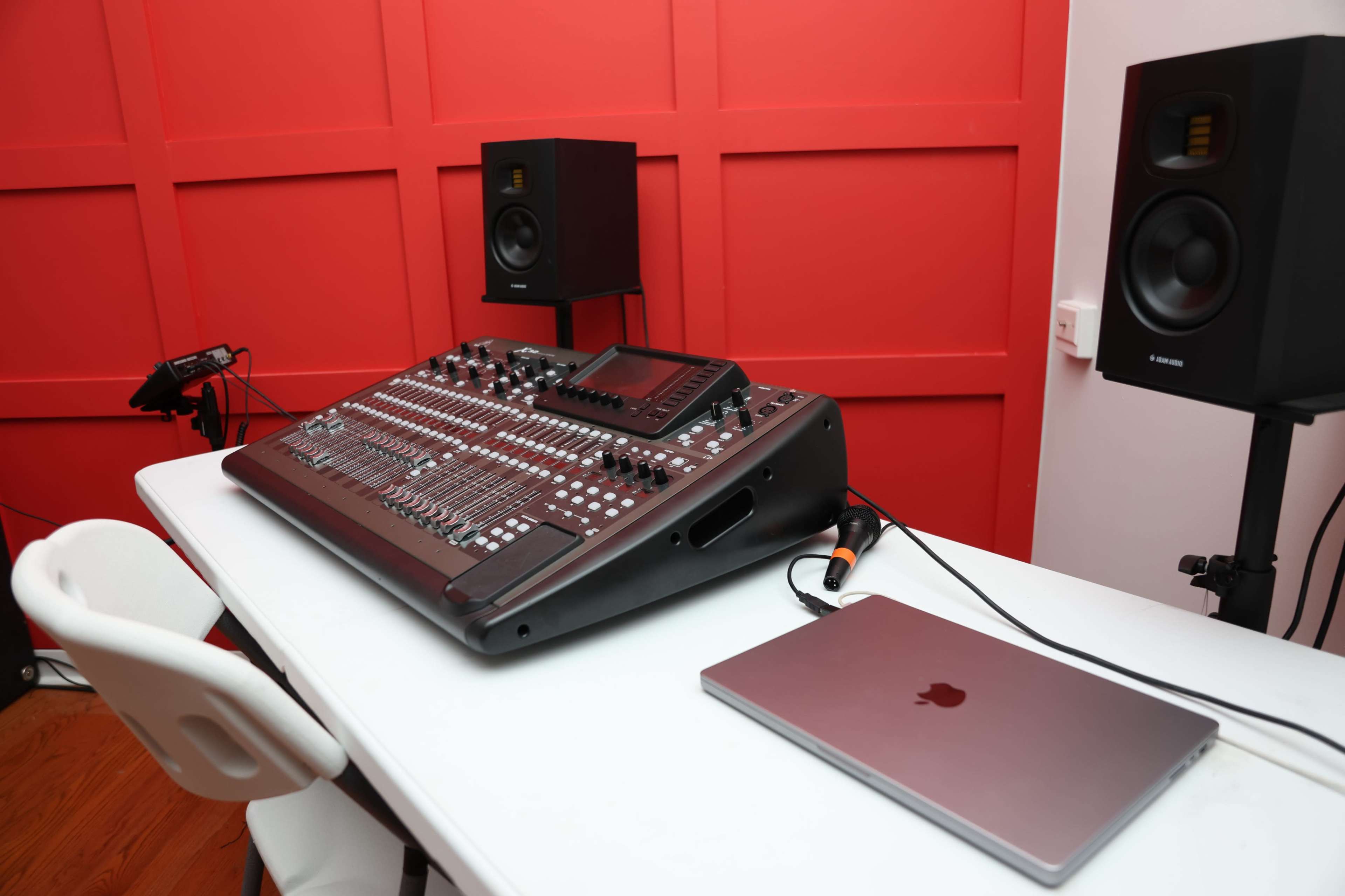 A mixing console is set on a white table beside a laptop, with black studio monitors positioned against a bright red wall.