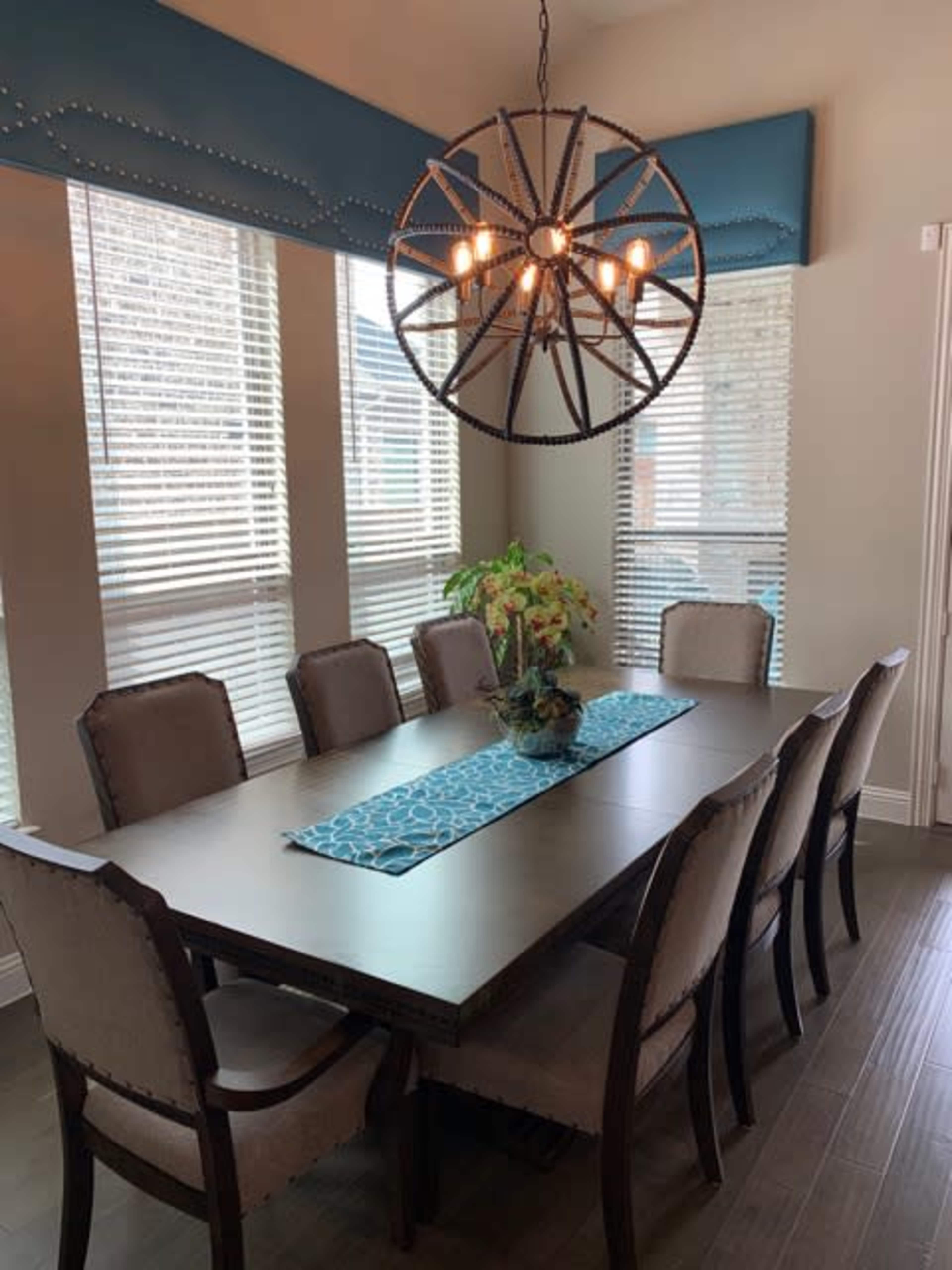 A large wooden dining table is set in a well-lit room with a chandelier above and a decorative centerpiece, surrounded by upholstered chairs.