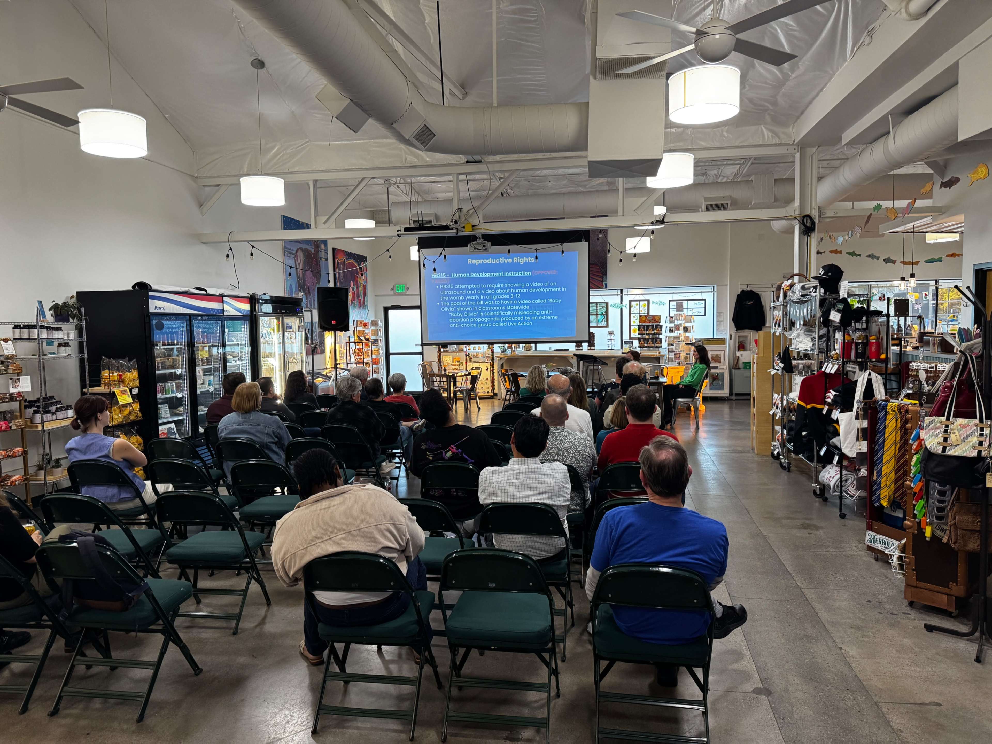 A group of people sits in folding chairs facing a large screen displaying information in a spacious indoor venue filled with shelves of merchandise.