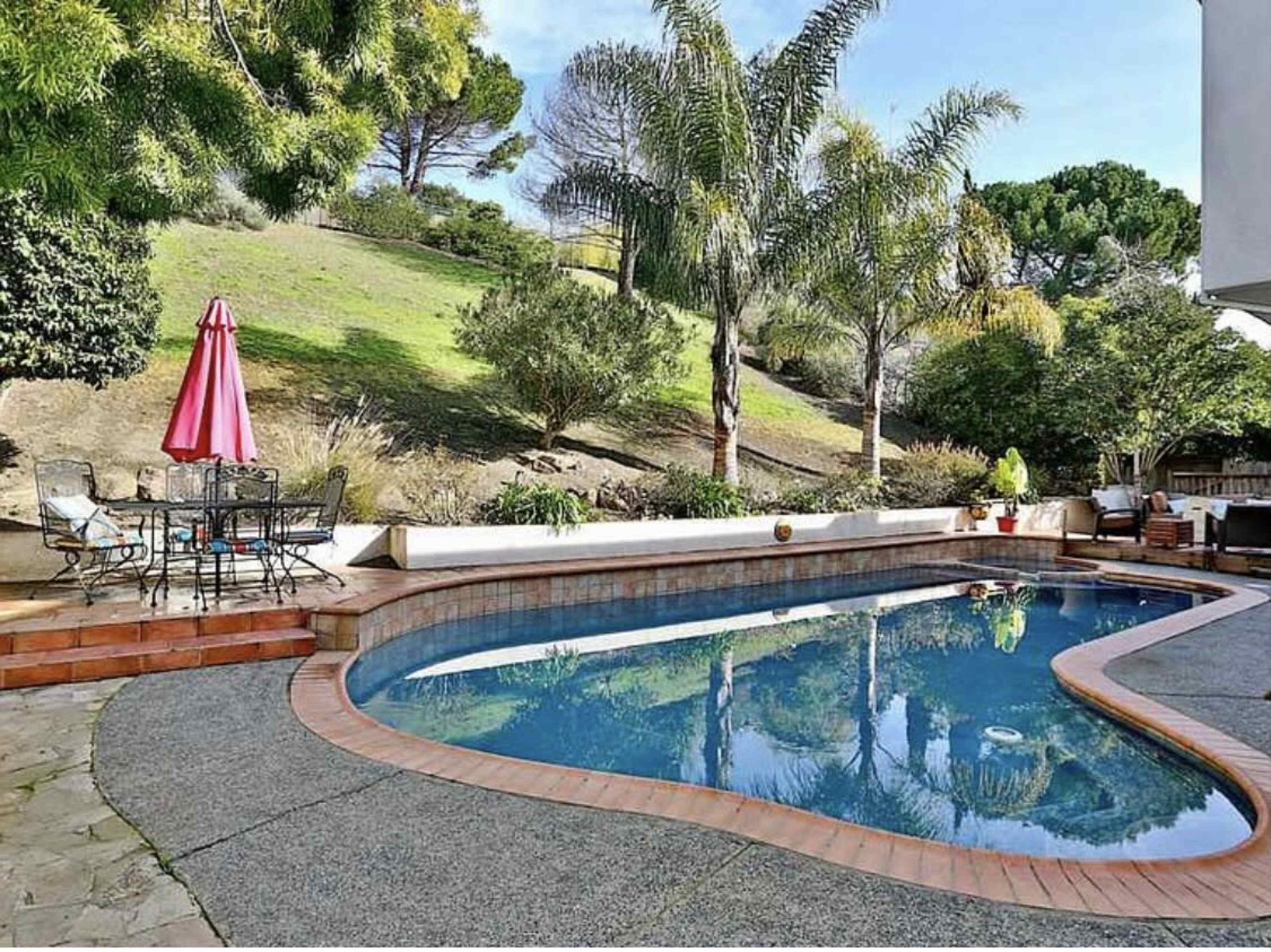 A landscaped backyard with a swimming pool surrounded by palm trees and outdoor seating under a pink umbrella.