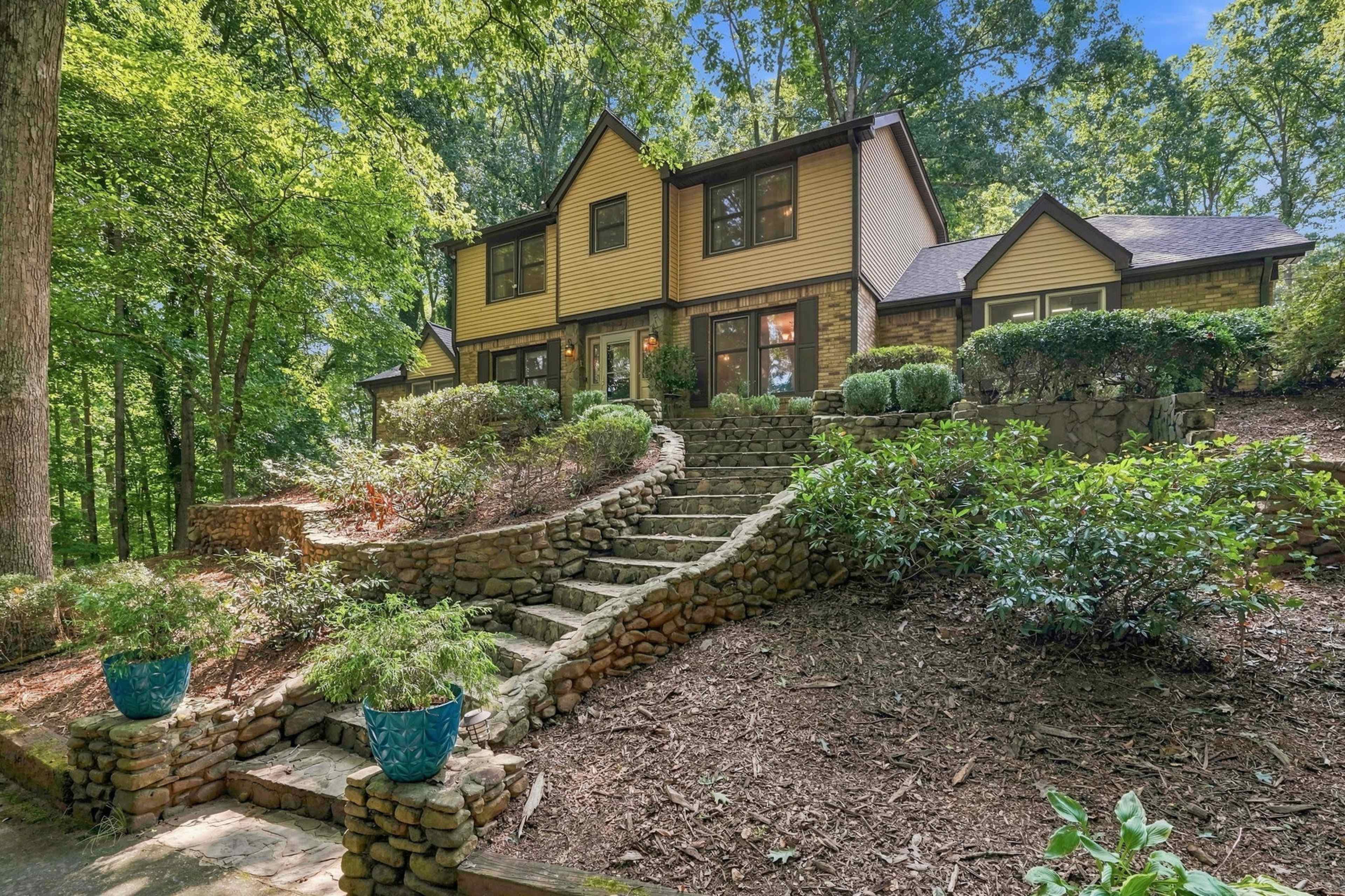 A two-story house with a mix of stone and wood siding is situated on a landscaped hill, featuring a stone pathway that leads to the entrance.