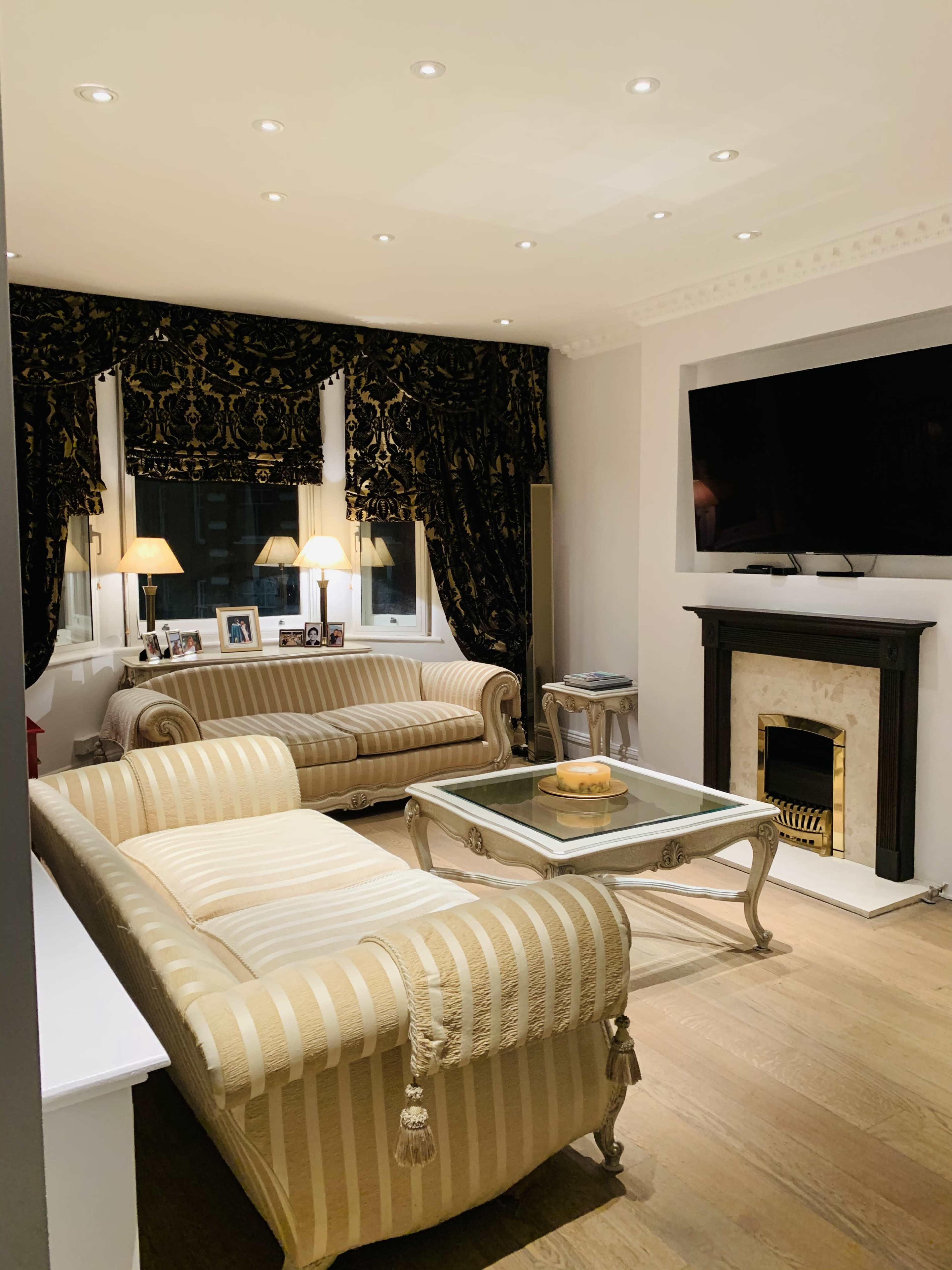 The living room features two striped sofas, a glass coffee table, and a TV mounted above a black marble fireplace, all illuminated by recessed ceiling lights.